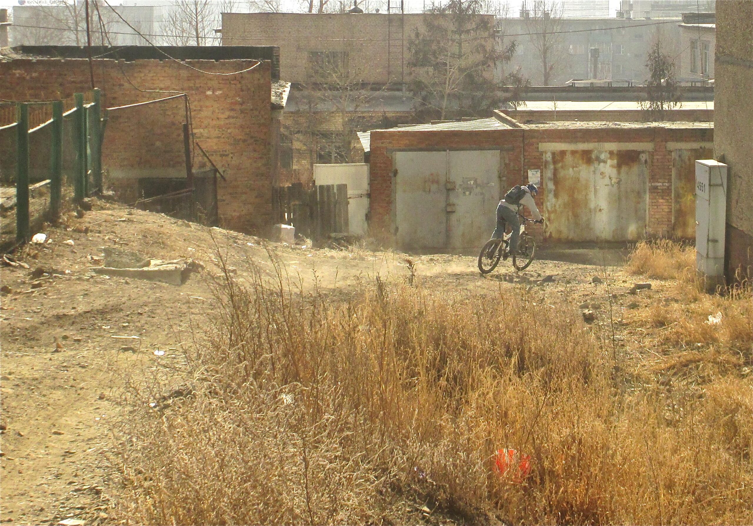 A person riding a mountain bike on a dirt path in an industrial area, surrounded by dry grass and buildings. The scene is dusty with a clear sky in the background. District 15 Network mountain bike trail.