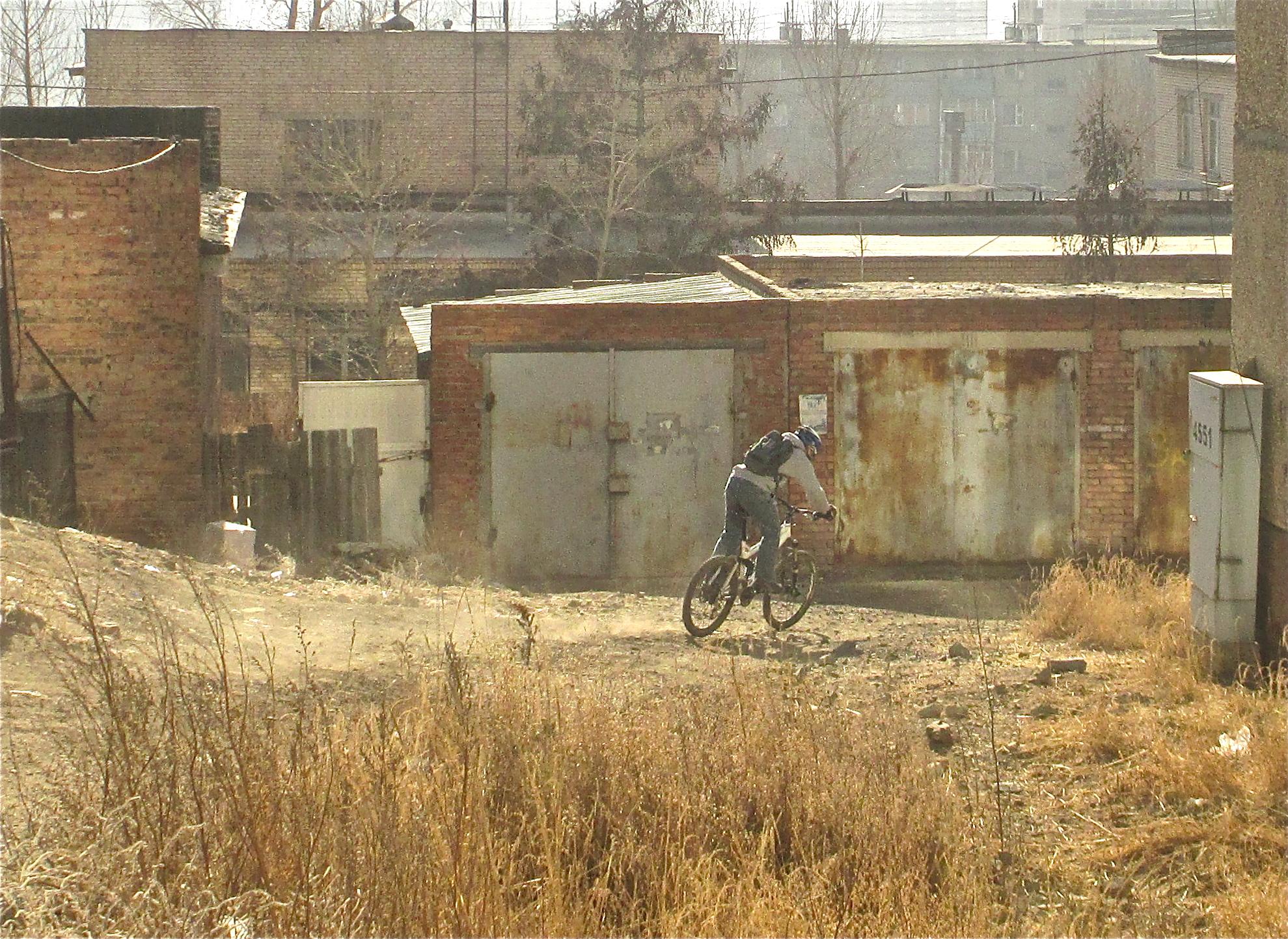 A mountain biker rides on a dirt path surrounded by overgrown grass and an industrial area, featuring weathered brick buildings and metal garages in the background. Dust is rising behind the bike as it navigates the terrain on a sunny day. District 15 Network mountain bike trail.