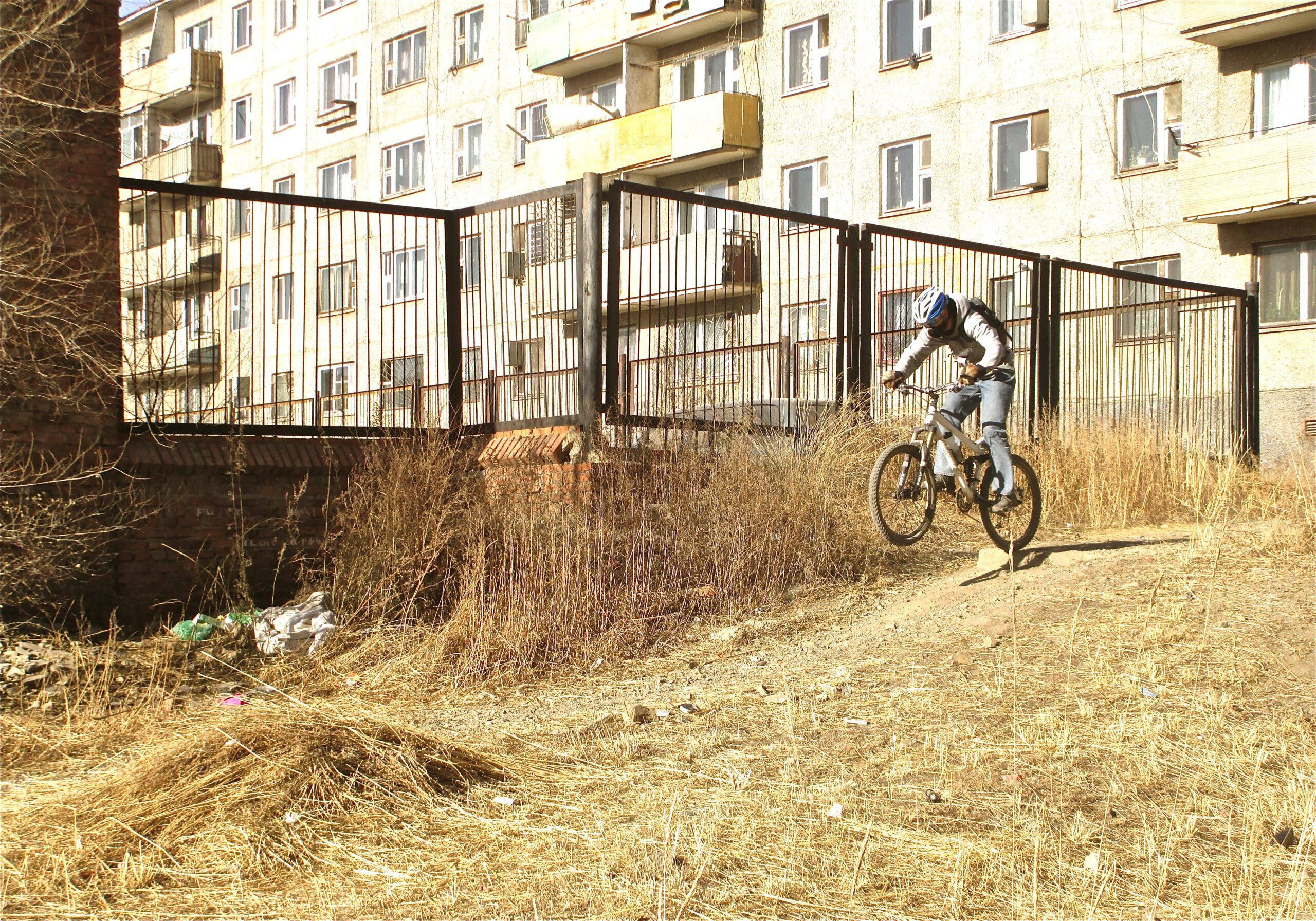 A cyclist performing a jump on a mountain bike in an outdoor area with dried grass and weeds, near a fenced structure and an apartment building in the background. District 15 Network mountain bike trail.