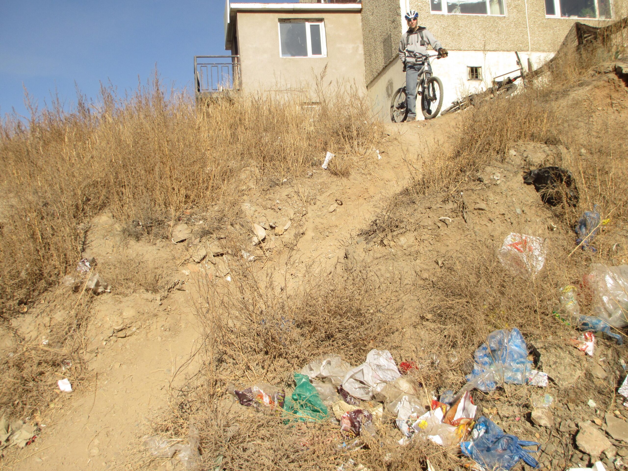 A cyclist in gray clothing and a helmet stands with a mountain bike on a dirt slope covered with dry grass and scattered litter. In the background, there is a two-story building with a balcony and large windows. The hillside has a steep incline with visible rocks and trash mixed in with the vegetation. The sky is clear and blue. District 15 Network mountain bike trail.
