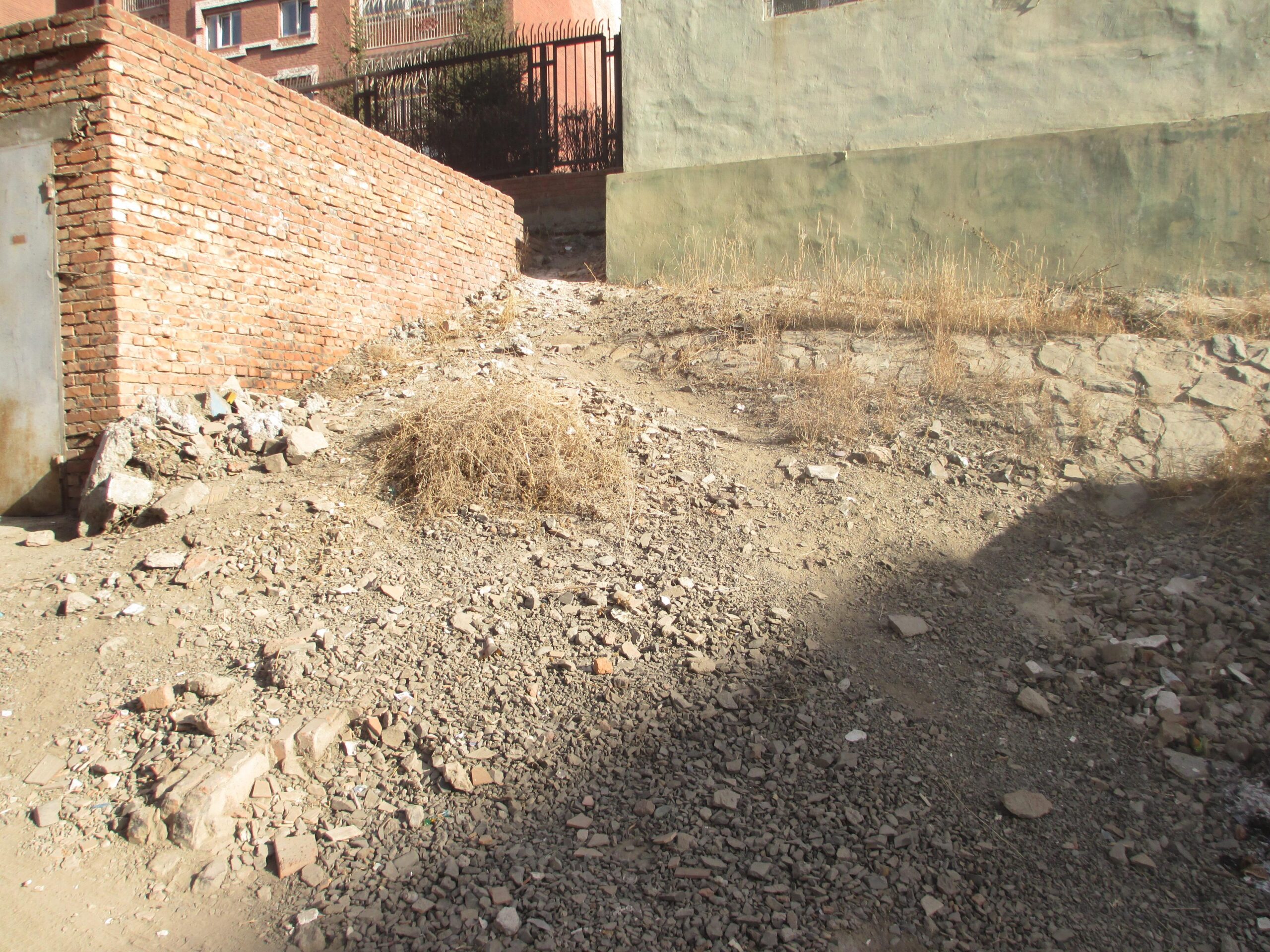 A gravelly pathway leading up a slope, bordered by a brick wall on the left and a green painted building on the right. The ground is uneven and scattered with small rocks and dried vegetation. Sunlight casts shadows across the terrain. District 15 Network mountain bike trail.
