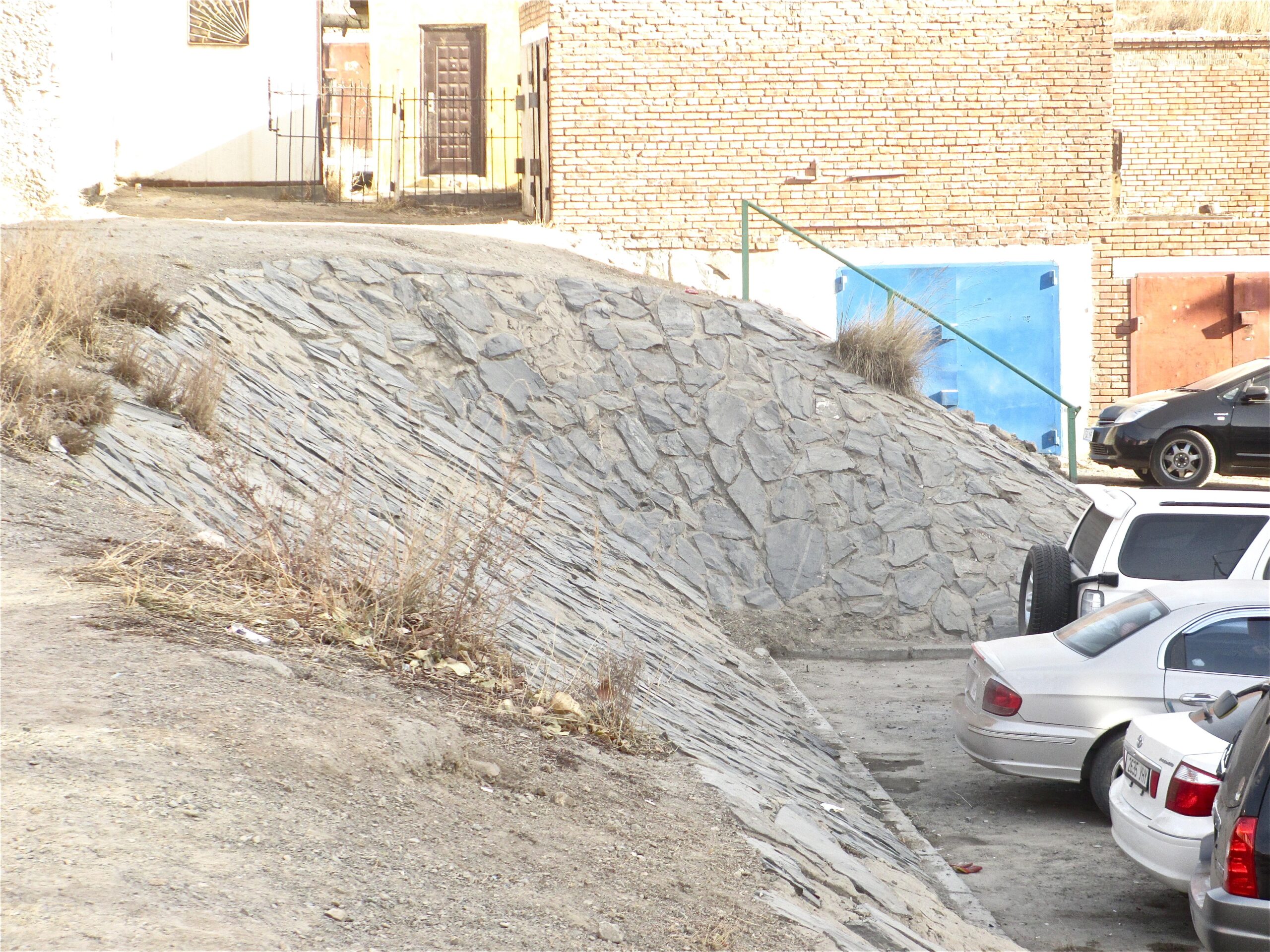 A sloped stone surface leading to a gravel area with parked cars. In the background, there are brick walls and a set of metal doors, along with a gated pathway. Dry grass and debris are visible along the edges of the slope. District 15 Network mountain bike trail.