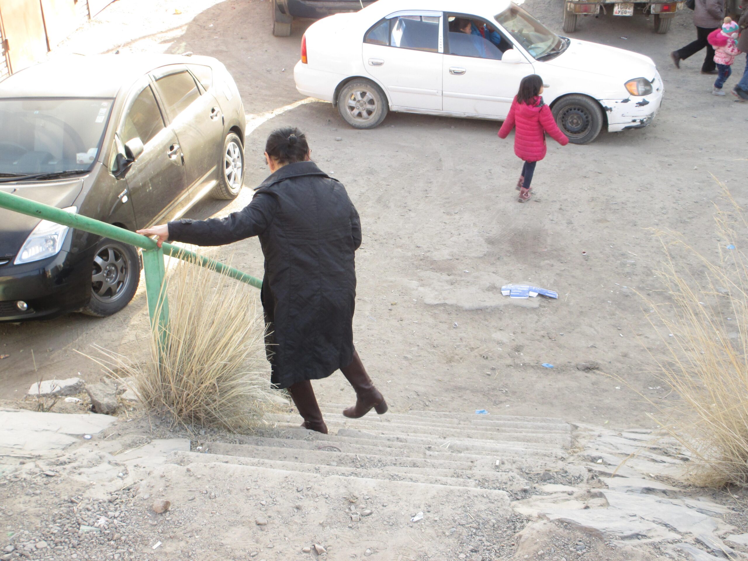 A woman in a black coat descends a set of stairs while holding onto a green railing. In the background, several parked cars are visible on a dirt road, and a girl in a pink jacket walks past a white car. The scene reflects an urban environment with sparse vegetation and scattered debris. District 15 Network mountain bike trail.