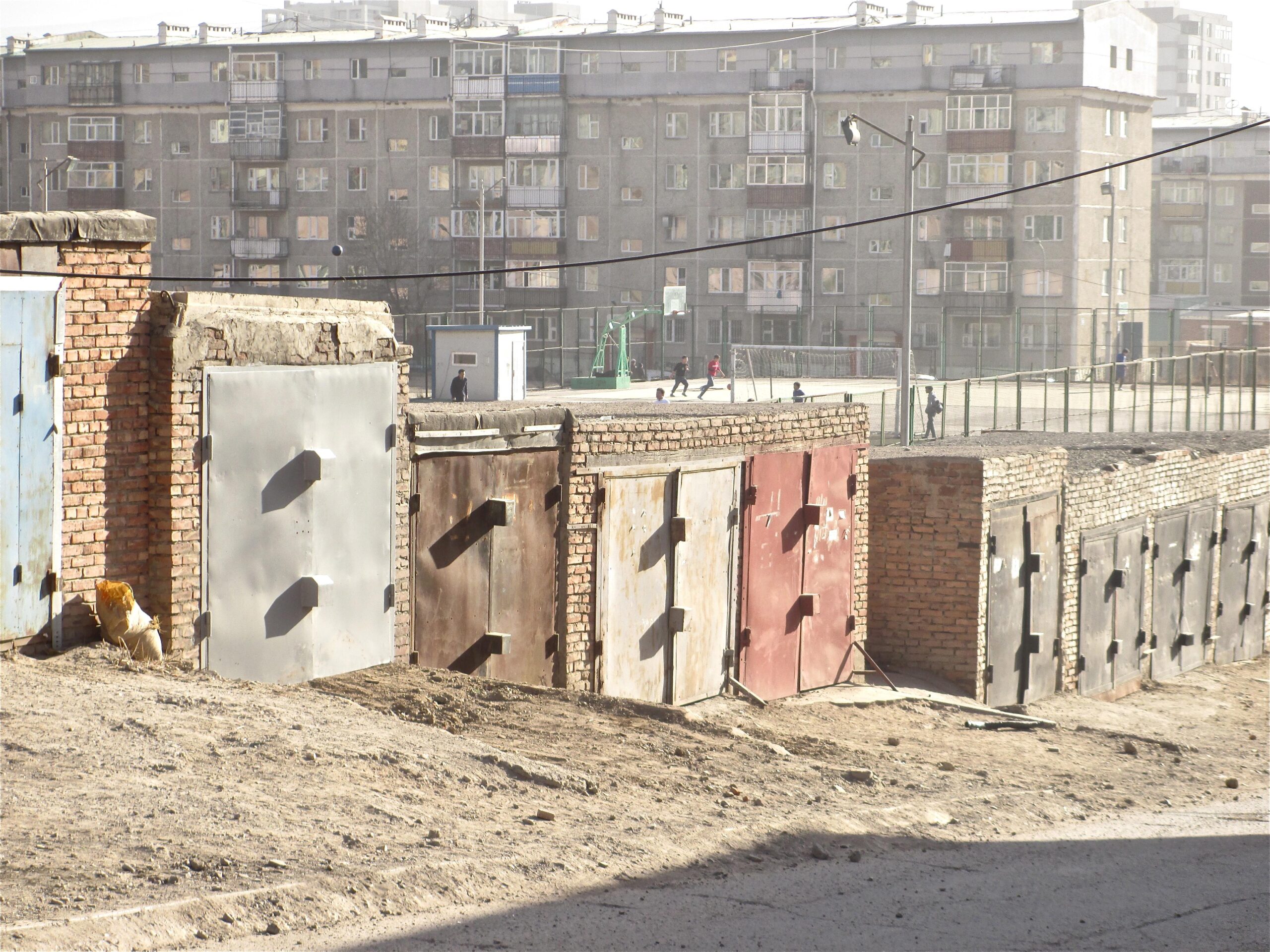 A row of brick storage facilities with metal doors, set against a backdrop of a multi-story residential building. In the distance, a group of children is playing on a sports field, with a fence enclosing the area. The scene is illuminated by daylight, giving a gritty urban feel. District 15 Network mountain bike trail.