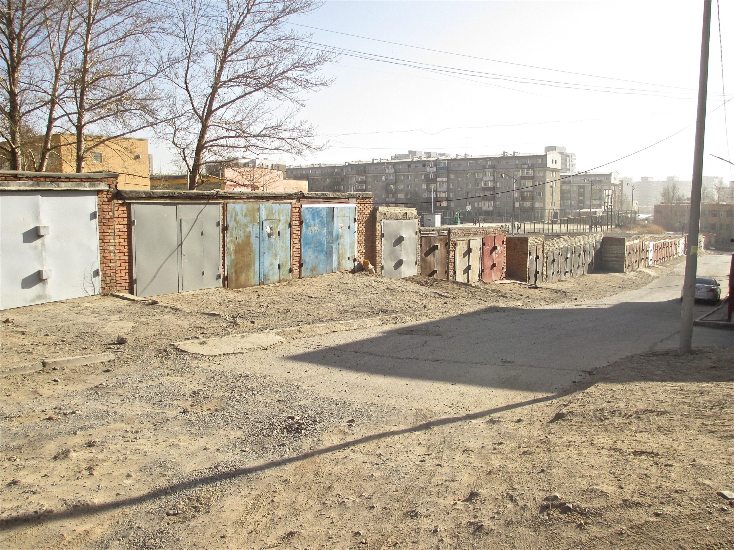 A narrow, dirt road lined with garages made of brick and metal, some showing signs of rust and wear. In the background, several apartment buildings can be seen under a clear sky. Barren trees are present, and utility poles run along the sides of the road. The scene reflects an urban environment with a somewhat desolate atmosphere. District 15 Network mountain bike trail.