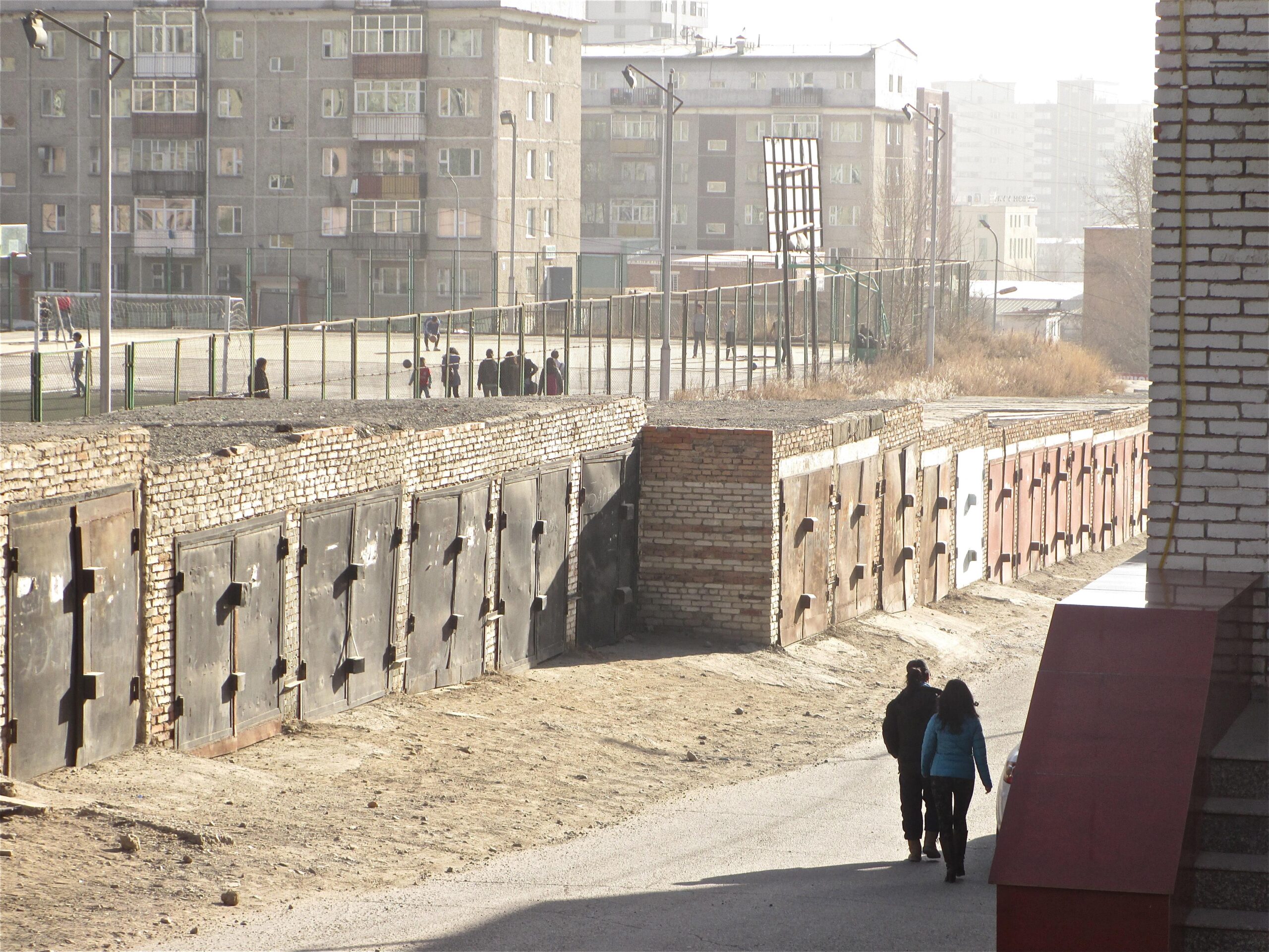 Two people walking along a dirt path beside brick garages with metal doors, while a group of people in the distance is gathered on a sports field. The background features residential buildings and a hazy atmosphere. District 15 Network mountain bike trail.