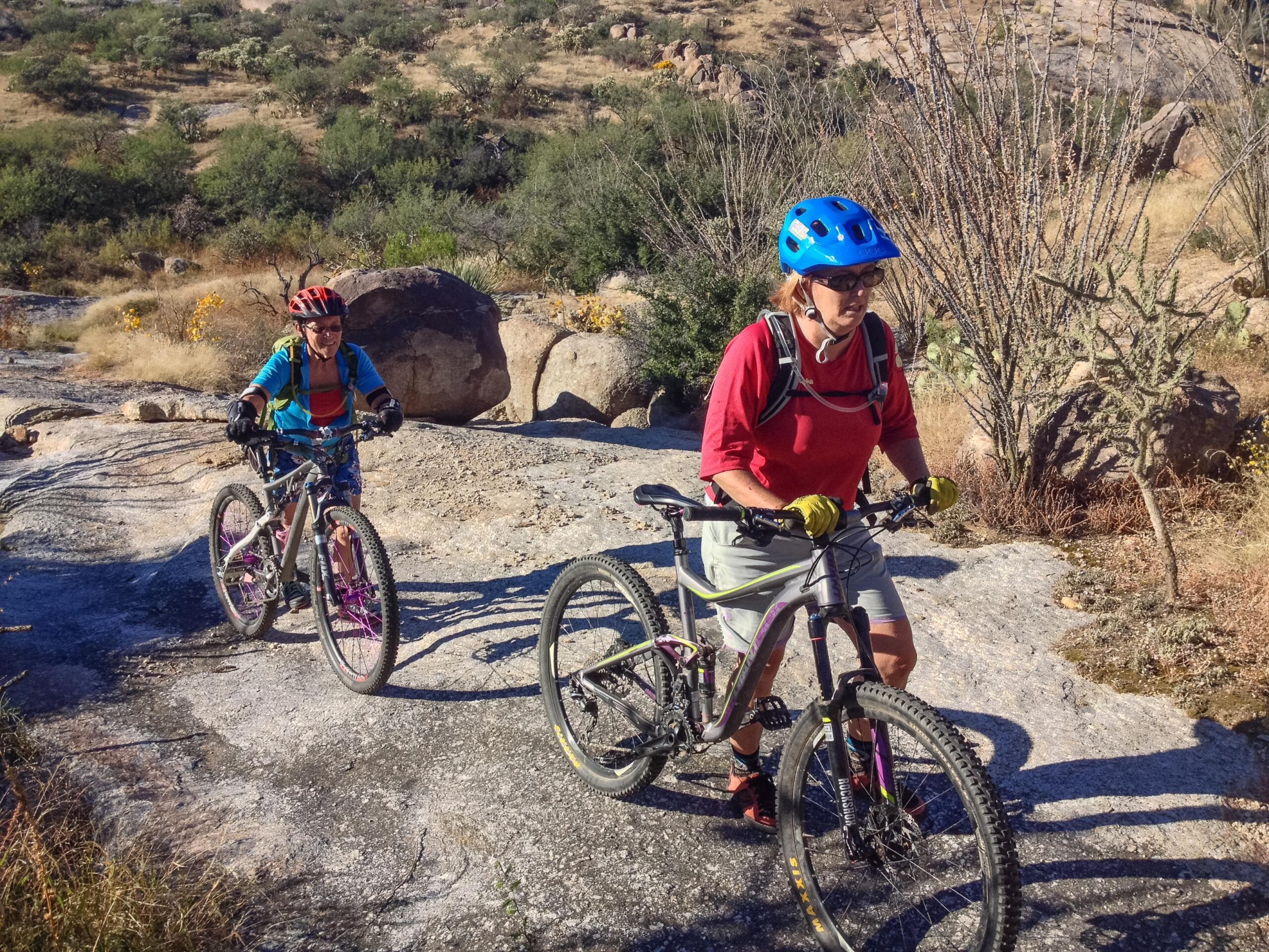 Two mountain bikers navigating a rocky trail in a desert landscape. One rider, wearing a bright blue helmet and a colorful jacket, is smiling while pedaling uphill. The other rider, dressed in a red shirt and shorts, is walking her bike beside her. The scenery includes boulders and sparse vegetation typical of a rugged outdoor environment. 50-year Trail / Golder Ranch mountain bike trail.