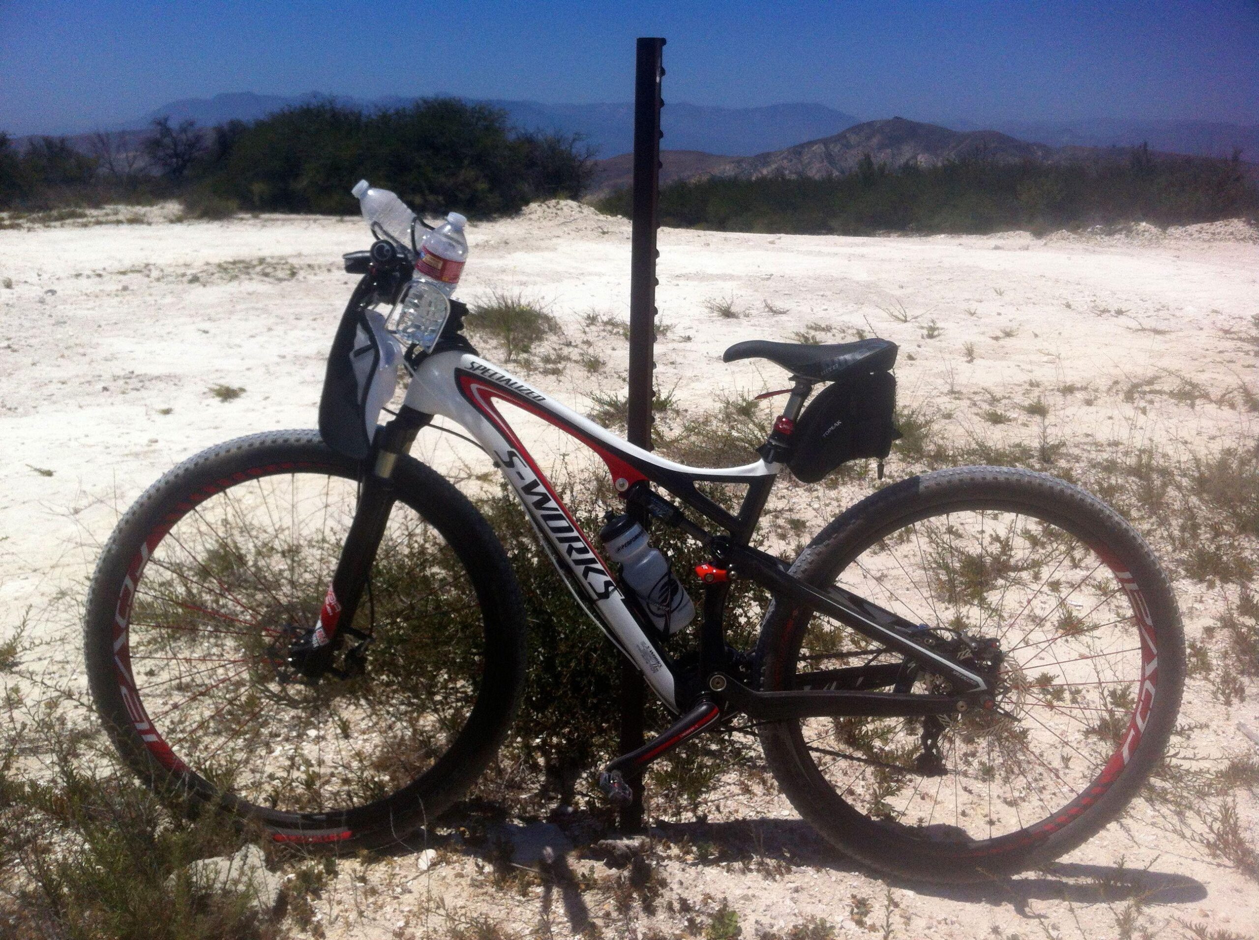 Specialized Epic: A mountain bike parked beside a metal post in a sandy landscape with sparse vegetation, under a clear blue sky. The bike features a white and black frame with red accents and is equipped with two water bottles attached to the frame. Distant hills can be seen in the background.