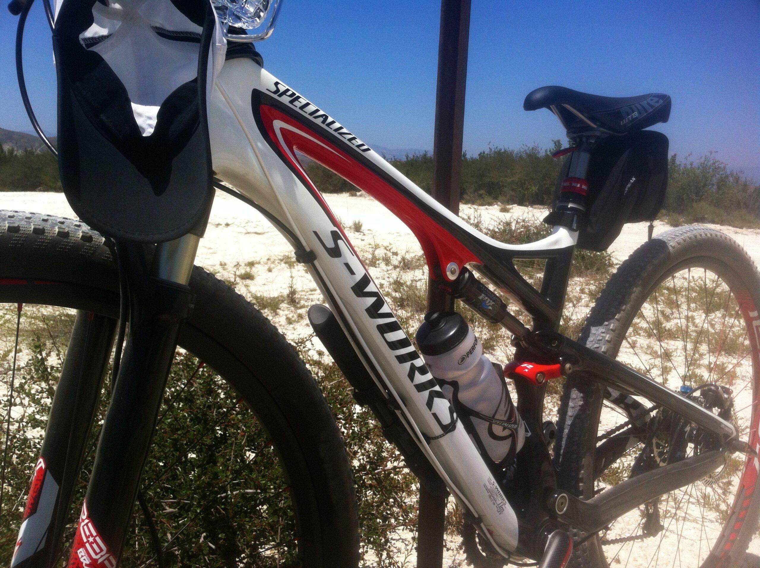 Specialized Epic: A close-up of a high-performance mountain bike, featuring a white and red frame labeled "S-Works." The bike includes a water bottle mounted on the frame and is positioned against a background of sandy terrain and sparse vegetation under a clear blue sky. The front suspension fork and tire are also visible, showcasing the bike's design and features.