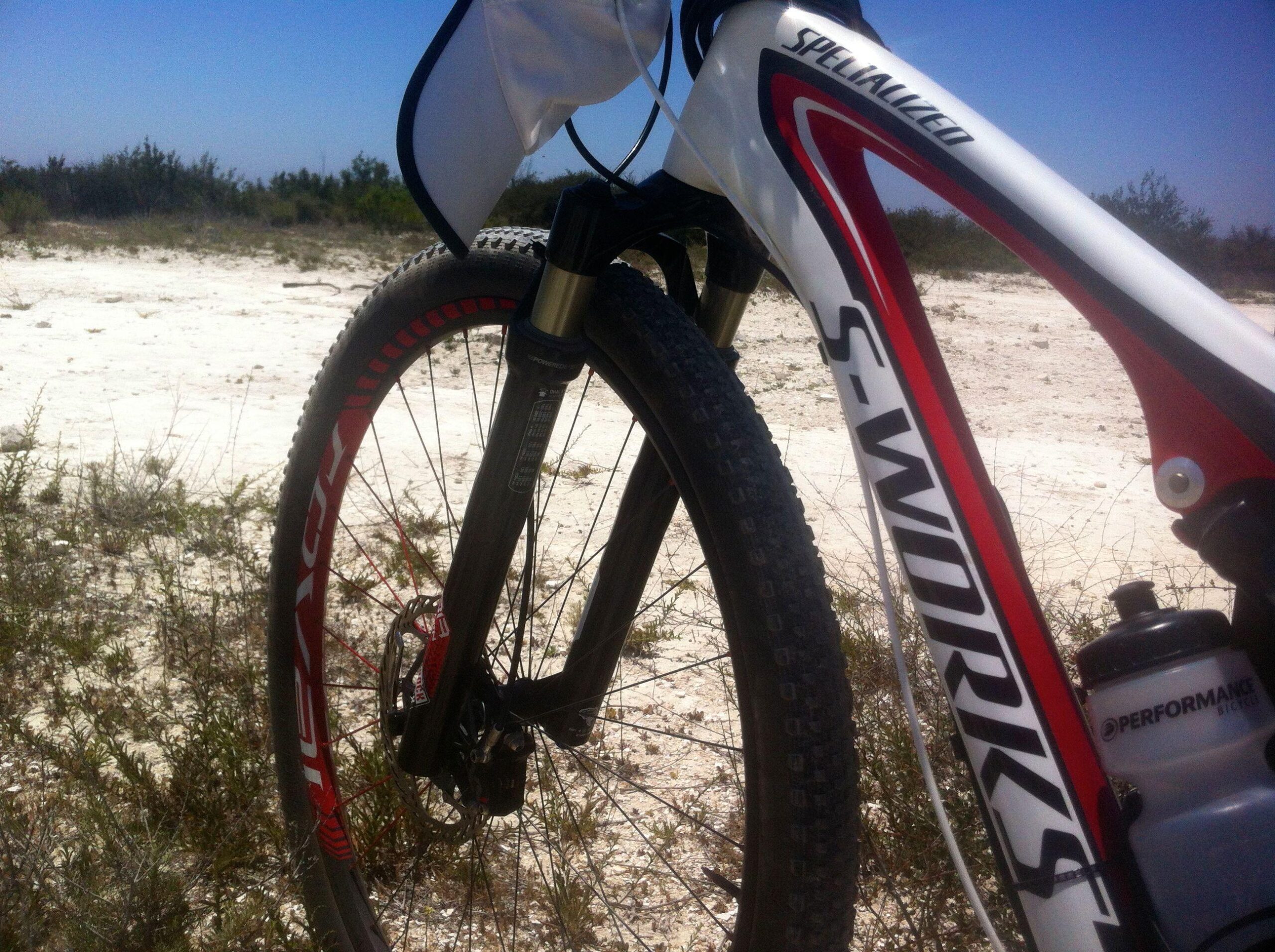 Specialized Epic: Close-up view of the front wheel and frame of a Specialized S-Works mountain bike, set against a sandy, open terrain with sparse vegetation. The bike features a black suspension fork, red and black tire, and a water bottle attached to the frame. The sky is clear and blue in the background.