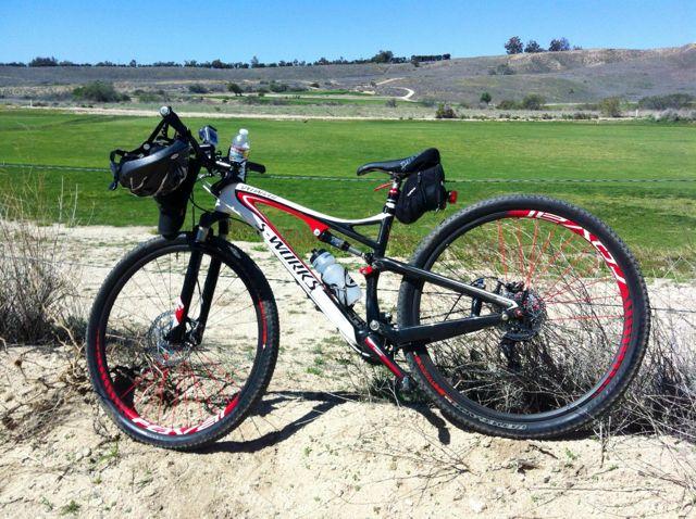 Specialized Epic: A mountain bike leaning against a bush on a sandy surface, set against a backdrop of green fields and distant hills under a clear blue sky. The bike features distinctive red and black wheels and a small bag attached to the frame.