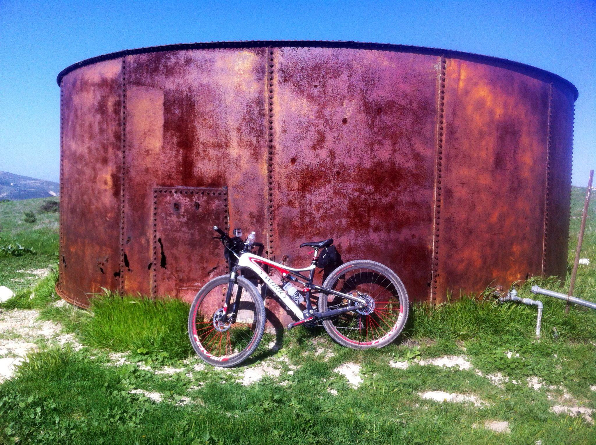 Specialized Epic: A mountain bike leaning against a rusted metal tank, situated on a grassy hillside under a clear blue sky.