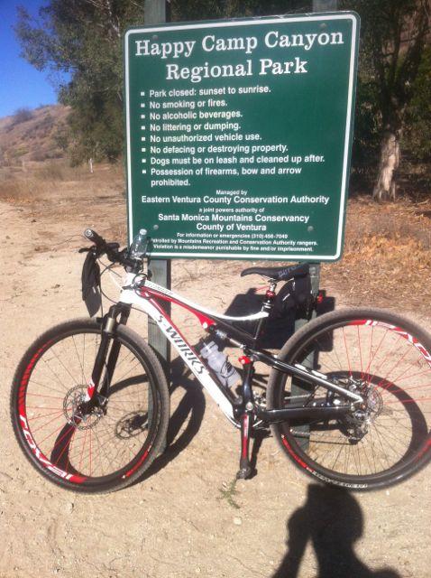 Specialized Epic: A mountain bike parked beside a sign for Happy Camp Canyon Regional Park, listing park rules such as no smoking, no alcohol, and no littering. The background features a dry, hilly landscape typical of a park setting.