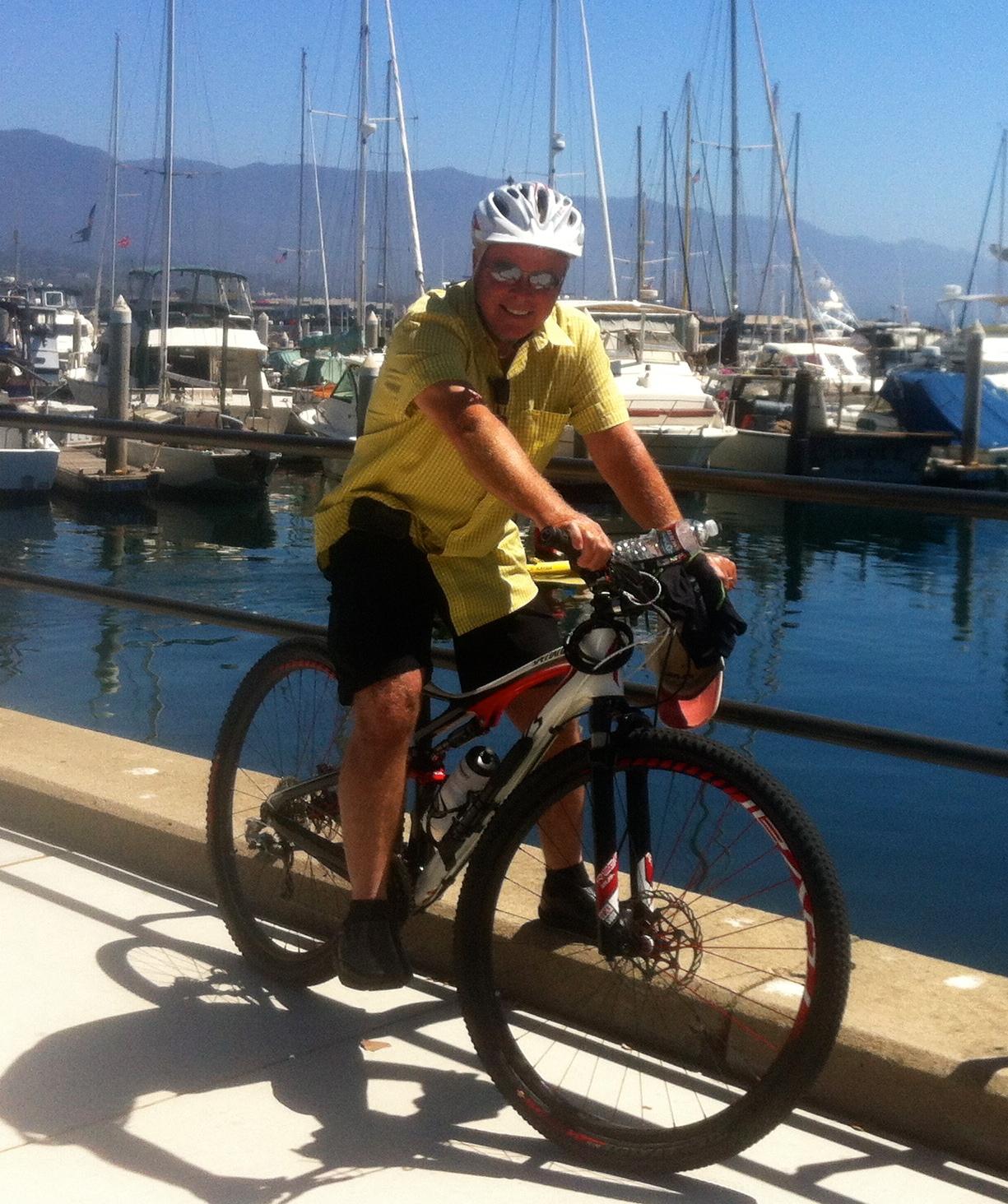 Specialized Epic: A smiling man wearing a yellow shirt and a white helmet is sitting on a mountain bike near a marina. In the background, there are several sailboats and yachts docked, and the water reflects the bright blue sky. The scene is sunny and captures a relaxed outdoor atmosphere.