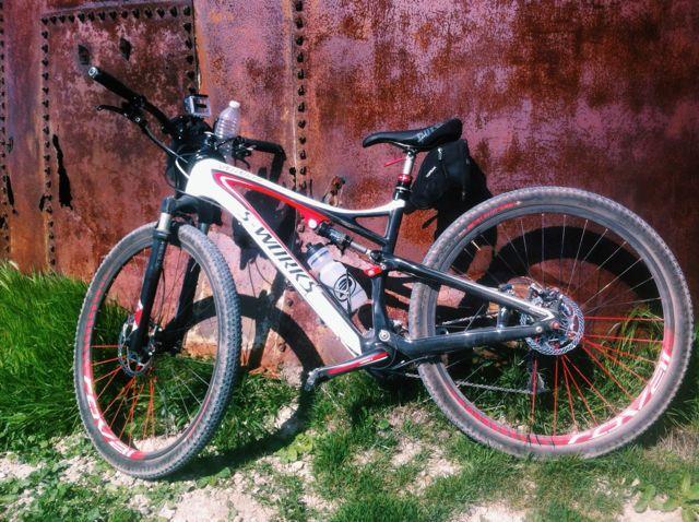 Specialized Epic: A mountain bike with red and white accents is leaning against a rusted metal structure. The bike has a water bottle mounted on the frame and a small bag attached to the seat. The background features green grass and a textured, weathered surface.