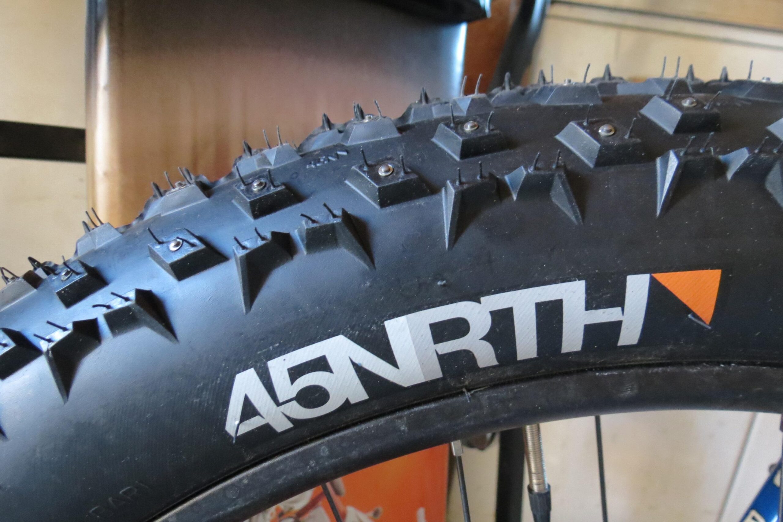 45Nrth Dillinger 4: Close-up image of a fat bike tire featuring sharp, pronounced treads and metal studs for enhanced grip on snow and ice. The tire is branded with the logo "45NRTH" in white letters, with an orange triangle. The background is slightly blurred, hinting at a garage or workshop setting.