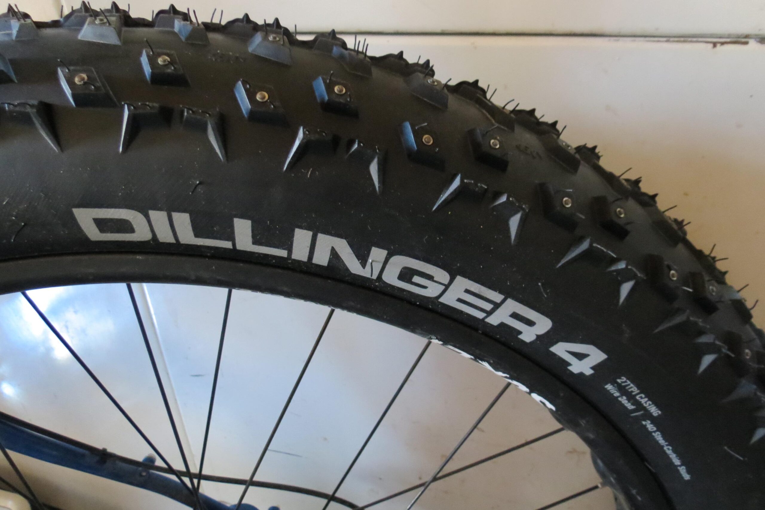 45Nrth Dillinger 4: Close-up image of a large, spiked fat bike tire labeled "DILLINGER 4." The tire features a rugged tread pattern designed for traction on snow and rough terrain. The background is a light surface, adding contrast to the dark tire.
