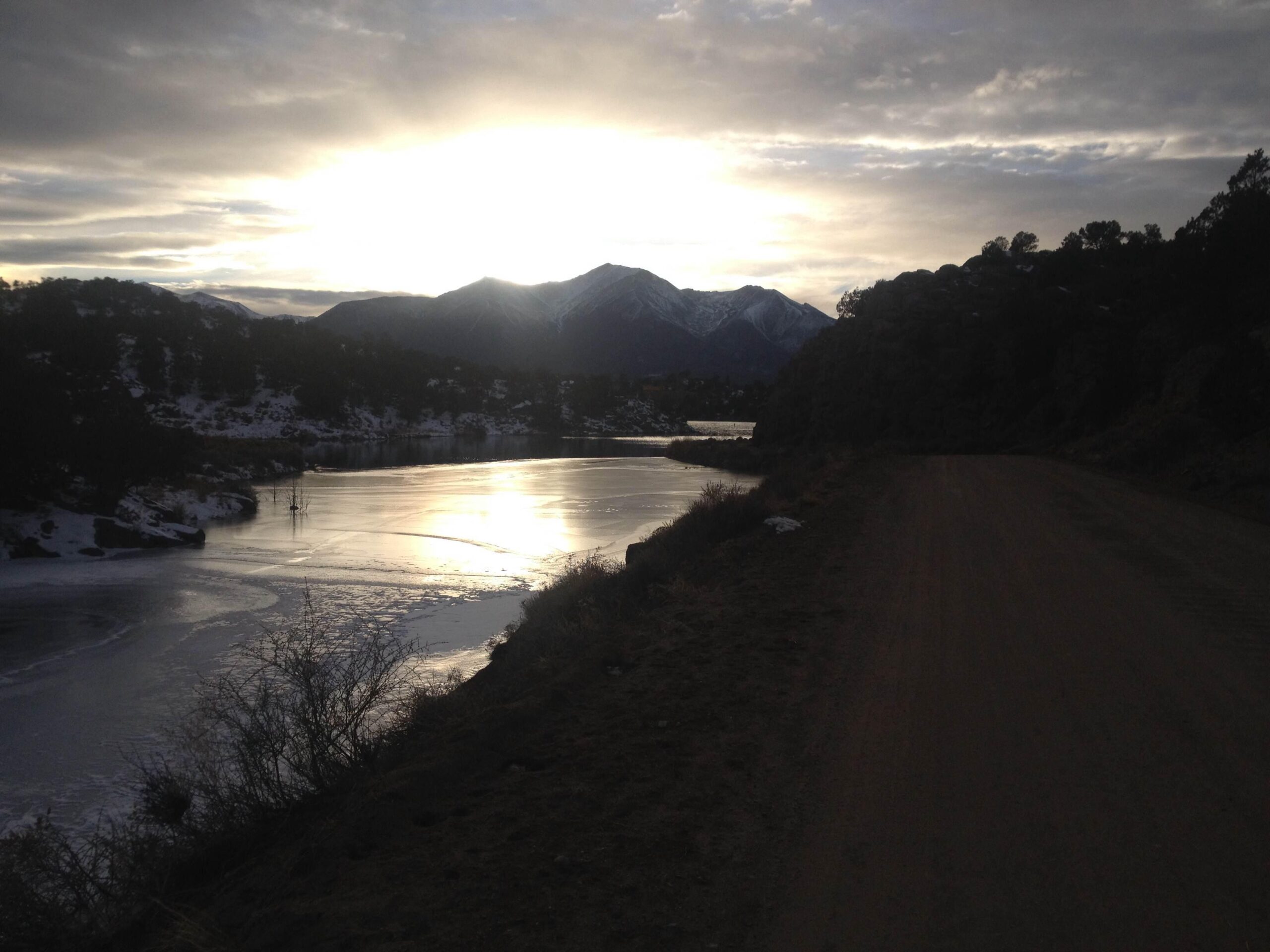 A serene landscape featuring a frozen river reflecting the soft glow of the sunrise. Snow-capped mountains line the background under a partly cloudy sky, while a dirt path runs alongside the riverbank, inviting exploration. Road #302 mountain bike trail.