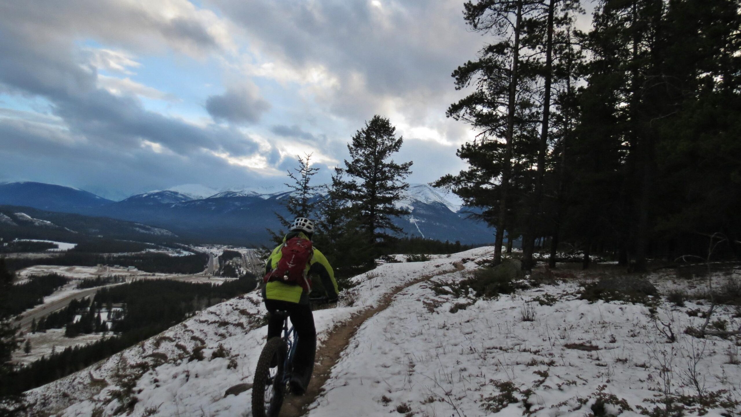 A cyclist in a bright yellow jacket rides a fat bike along a snowy trail in a mountainous landscape. Pine trees are visible on one side, while distant snow-capped mountains and a cloudy sky are seen in the background. Pyramid Bench mountain bike trail.