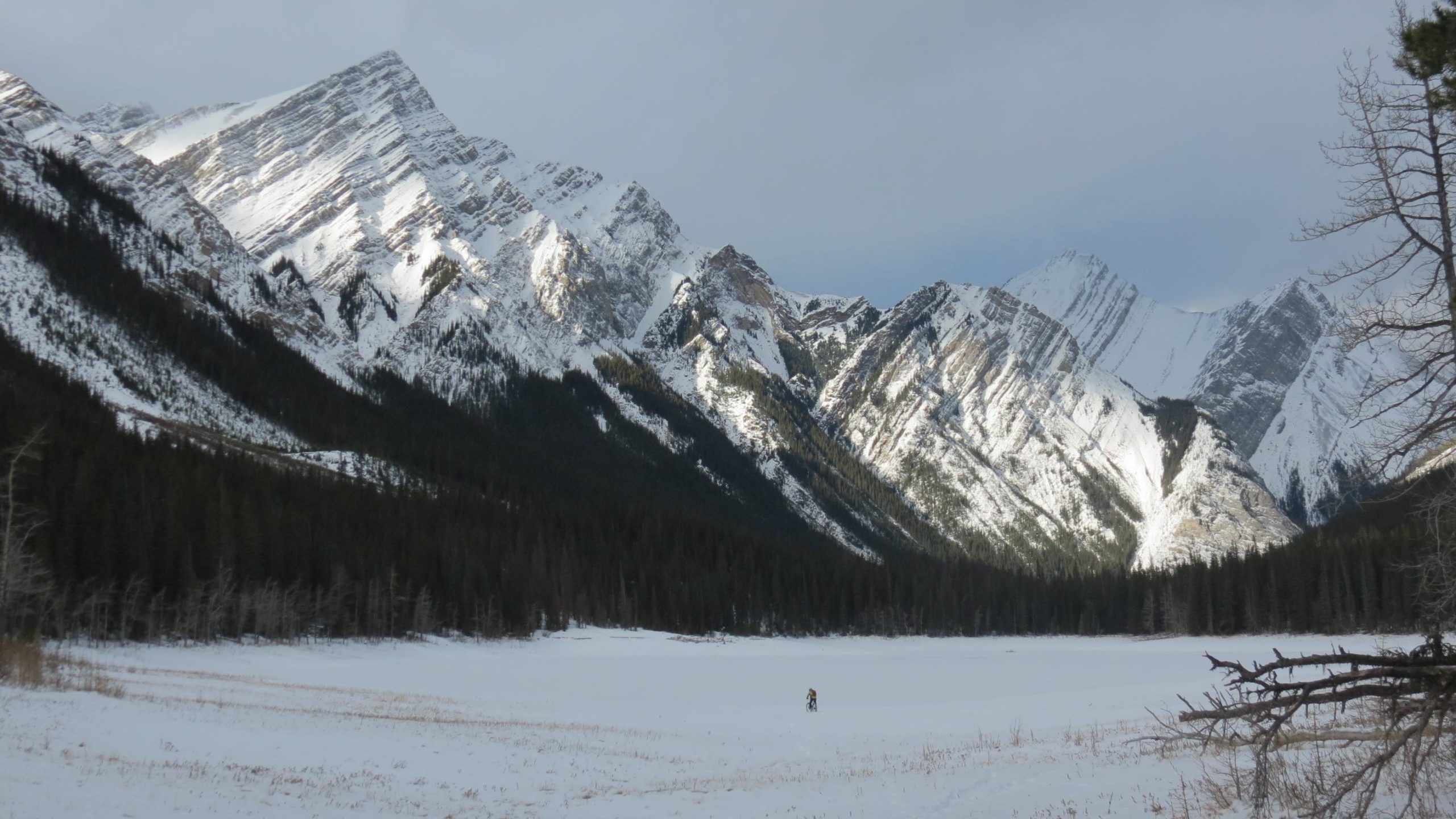 A serene winter landscape featuring towering snow-capped mountains in the background, with a patch of evergreen trees at the base. A vast, frozen expanse of snow-covered ground extends in the foreground, where a solitary figure is seen walking, creating a sense of scale against the grand natural scenery. The sky is cloudy, casting a soft light over the snowy scene. Jacques Lake mountain bike trail.