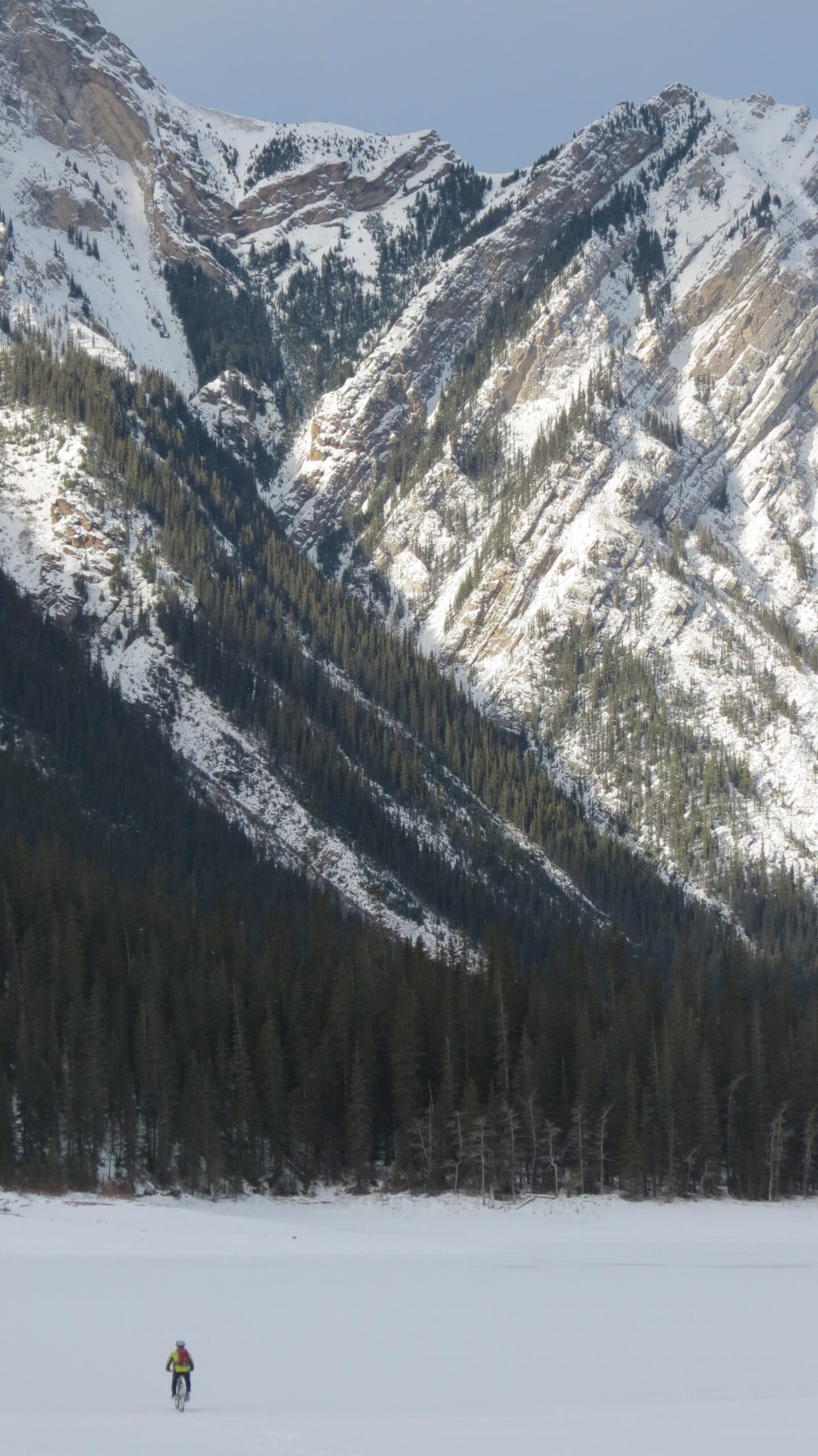 A cyclist riding on a snow-covered landscape with majestic snow-capped mountains and dense evergreen forests in the background. The scene showcases the beauty of winter outdoors. Jacques Lake mountain bike trail.