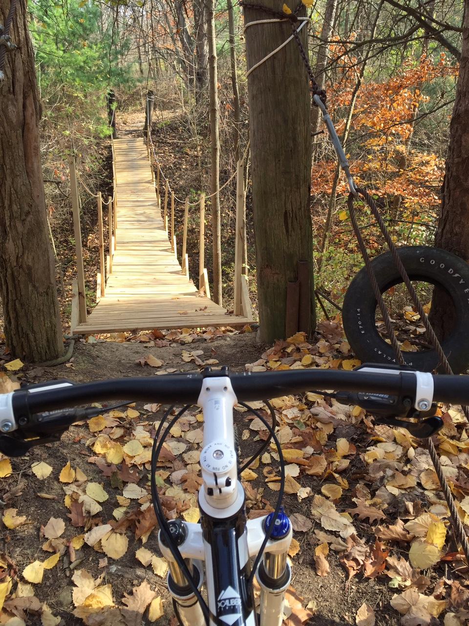 A view from the handlebars of a mountain bike, looking toward a wooden suspension bridge surrounded by autumn foliage. The ground is covered with fallen leaves, and a tire swing is visible to the side, suggesting a playful outdoor setting. Vultures Knob mountain bike trail.