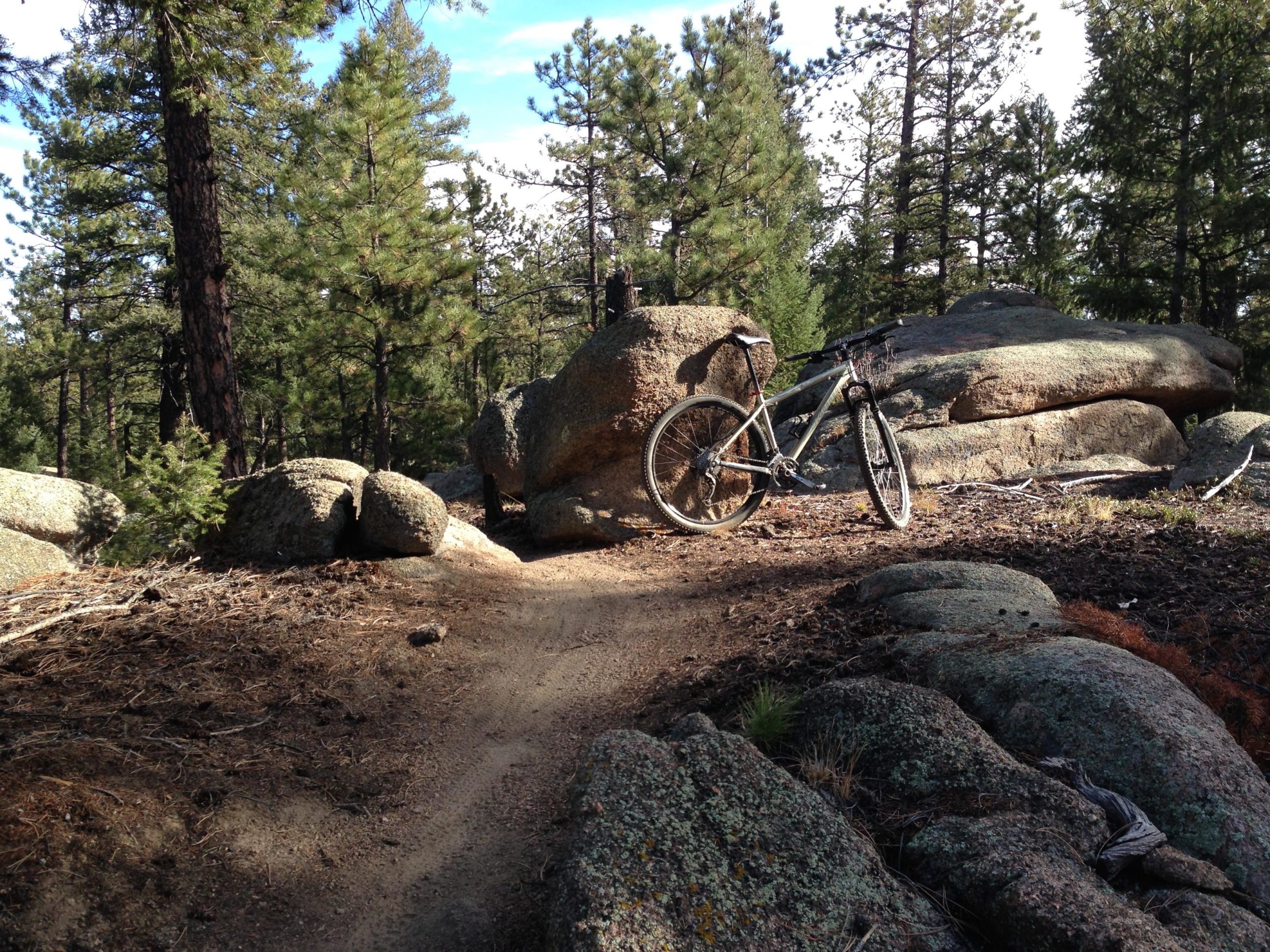 A mountain bike resting on a dirt trail surrounded by large boulders and tall pine trees in a forested setting. The sun is shining, illuminating the path and creating shadows from the rocks and bike. Little Scraggy mountain bike trail.