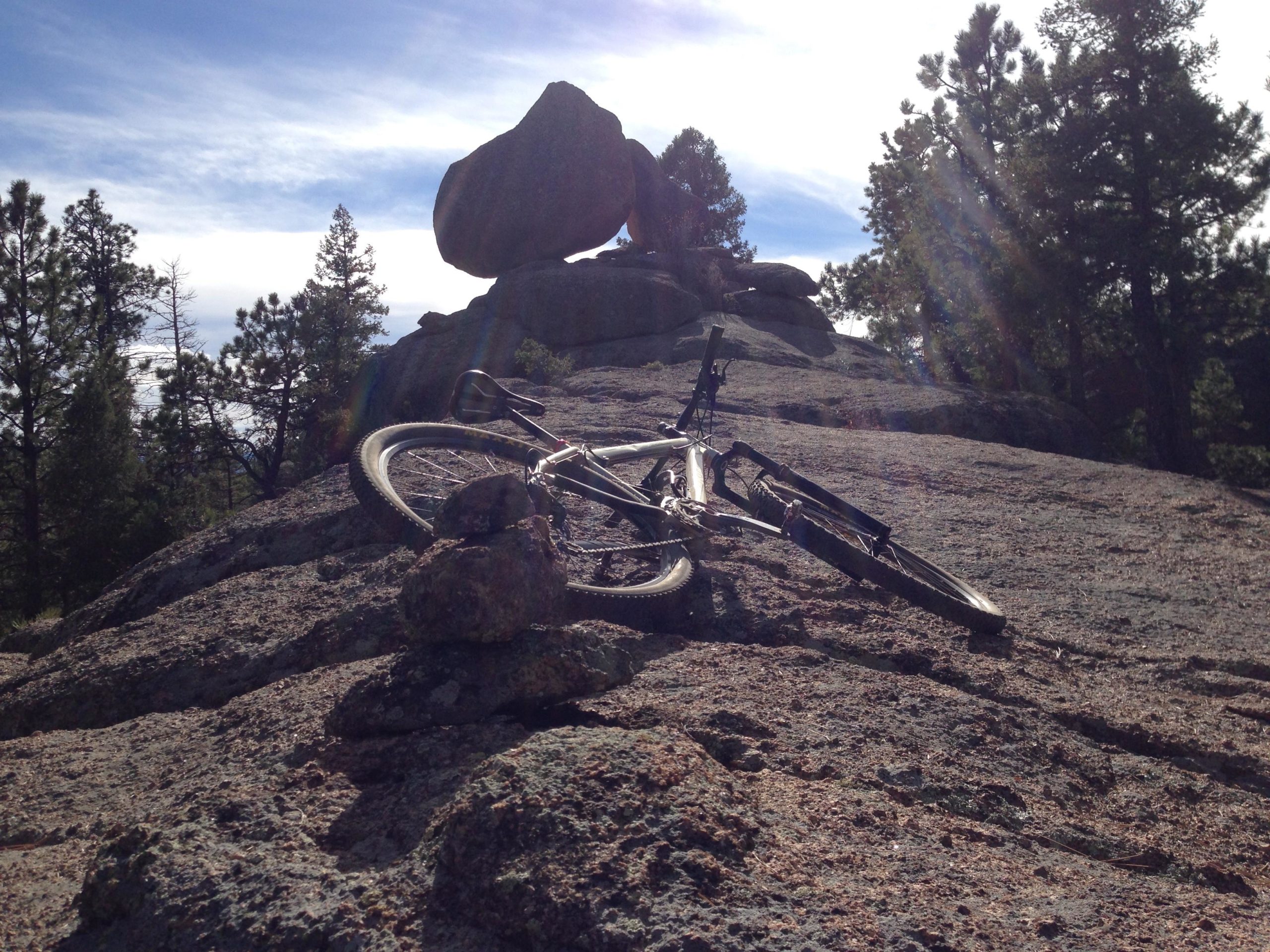 A mountain bike resting on rocky terrain, with a large boulder in the background and trees surrounding the area under a partly cloudy sky. Little Scraggy mountain bike trail.