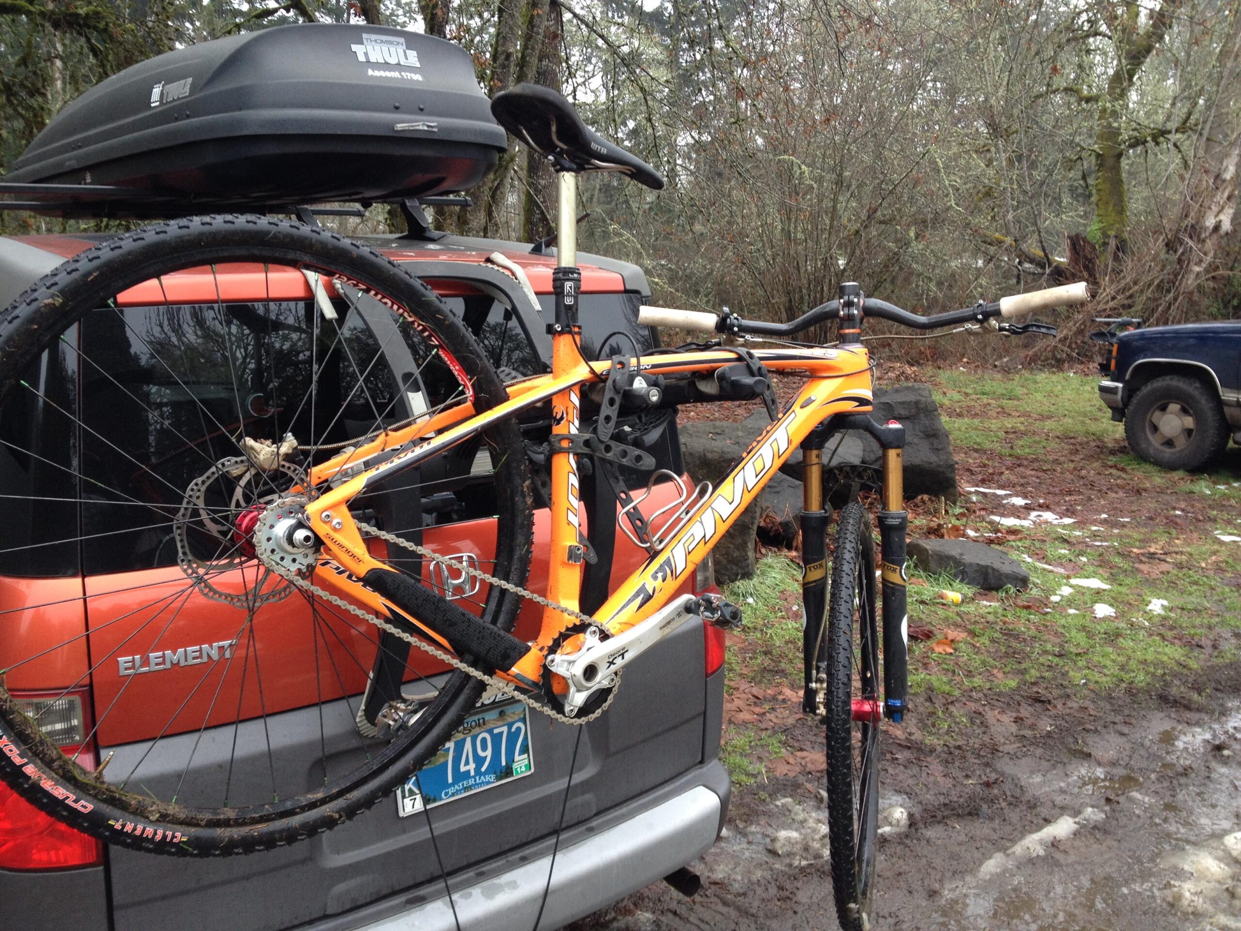 Pivot LES 29er: A bright orange mountain bike with a sleek frame is mounted on the back of a gray SUV. The bike is secured with a rack, showcasing its large tires and suspension. A black Thule cargo box is attached to the roof of the vehicle. The background features a wooded area with fallen leaves and rocky terrain, suggesting an outdoor setting.