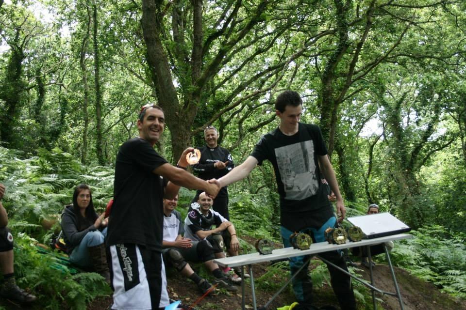 A group of people gathered in a forest, celebrating an event. Two individuals shake hands at the center; one is handing a trophy to the other. Several onlookers are seated in the background, smiling and watching the exchange. A table nearby displays more trophies. The scene is surrounded by lush greenery, highlighting a natural outdoor setting.