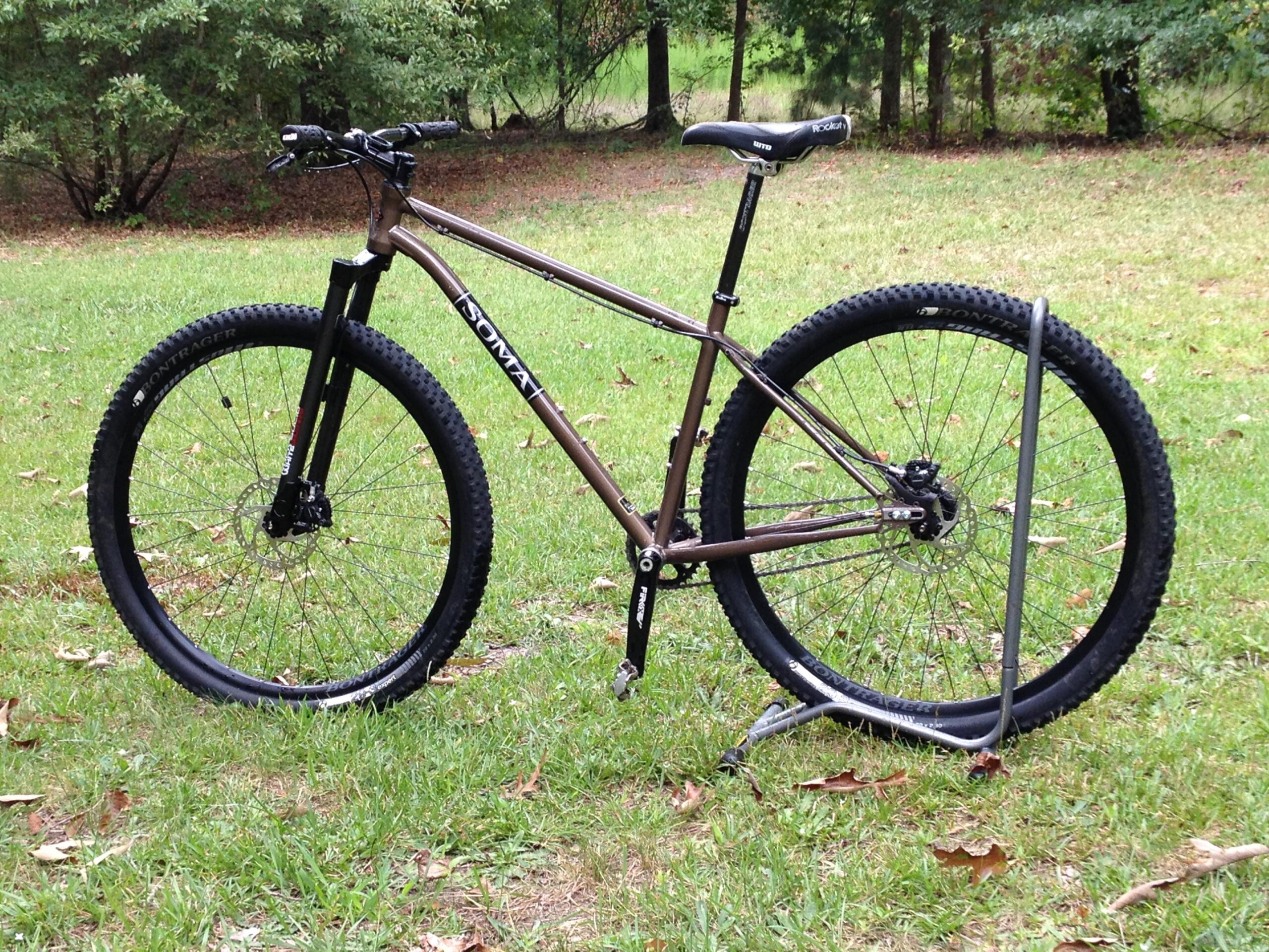 Soma Juice (Brownstone): A mountain bike with a sleek brown frame and wide, knobby tires is standing on green grass. The bike has a black front fork and disc brakes, showcasing its rugged build for off-road cycling. In the background, there are trees indicating a natural outdoor setting. A metal bike stand is partially visible, supporting the rear wheel.