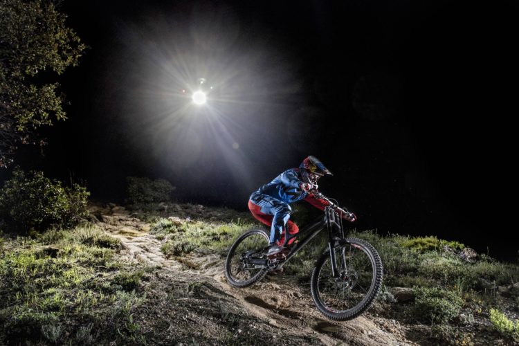A mountain biker in a blue and red outfit rides down a steep, rugged trail at night, illuminated by a bright light source above. The surrounding area features lush vegetation and rocky terrain, enhancing the dynamic movement of the biker.