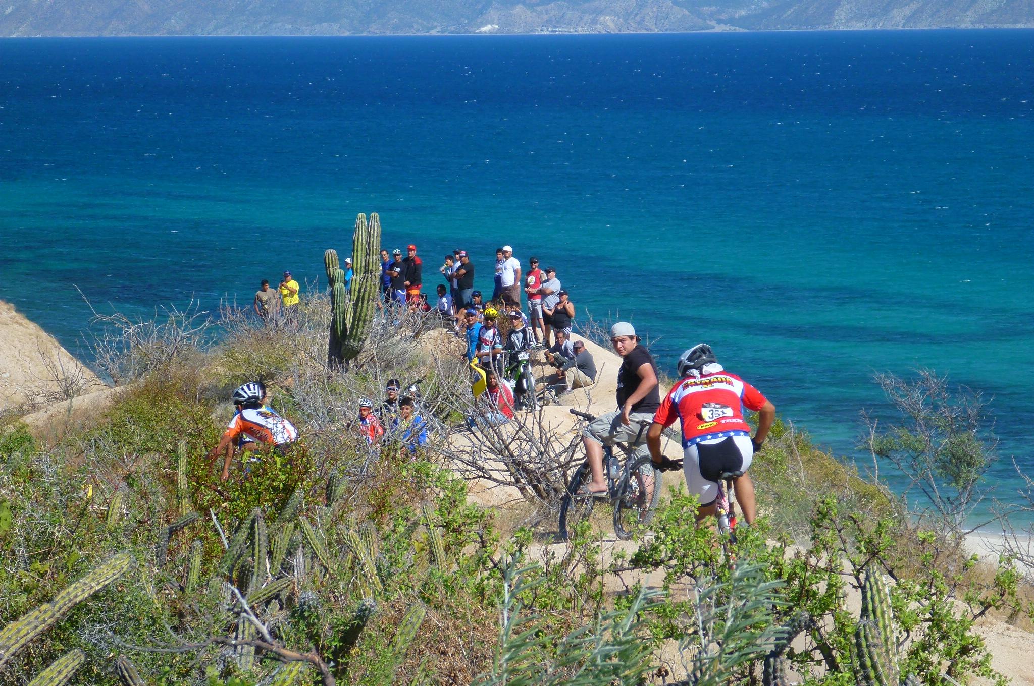 A scenic view of a coastal biking trail with several cyclists navigating the rocky terrain. In the background, a group of onlookers is gathered near the edge of the trail, enjoying the striking blue ocean and mountains in the distance. Cacti and other vegetation are visible along the path, adding to the natural landscape. Warren Ave mountain bike trail.