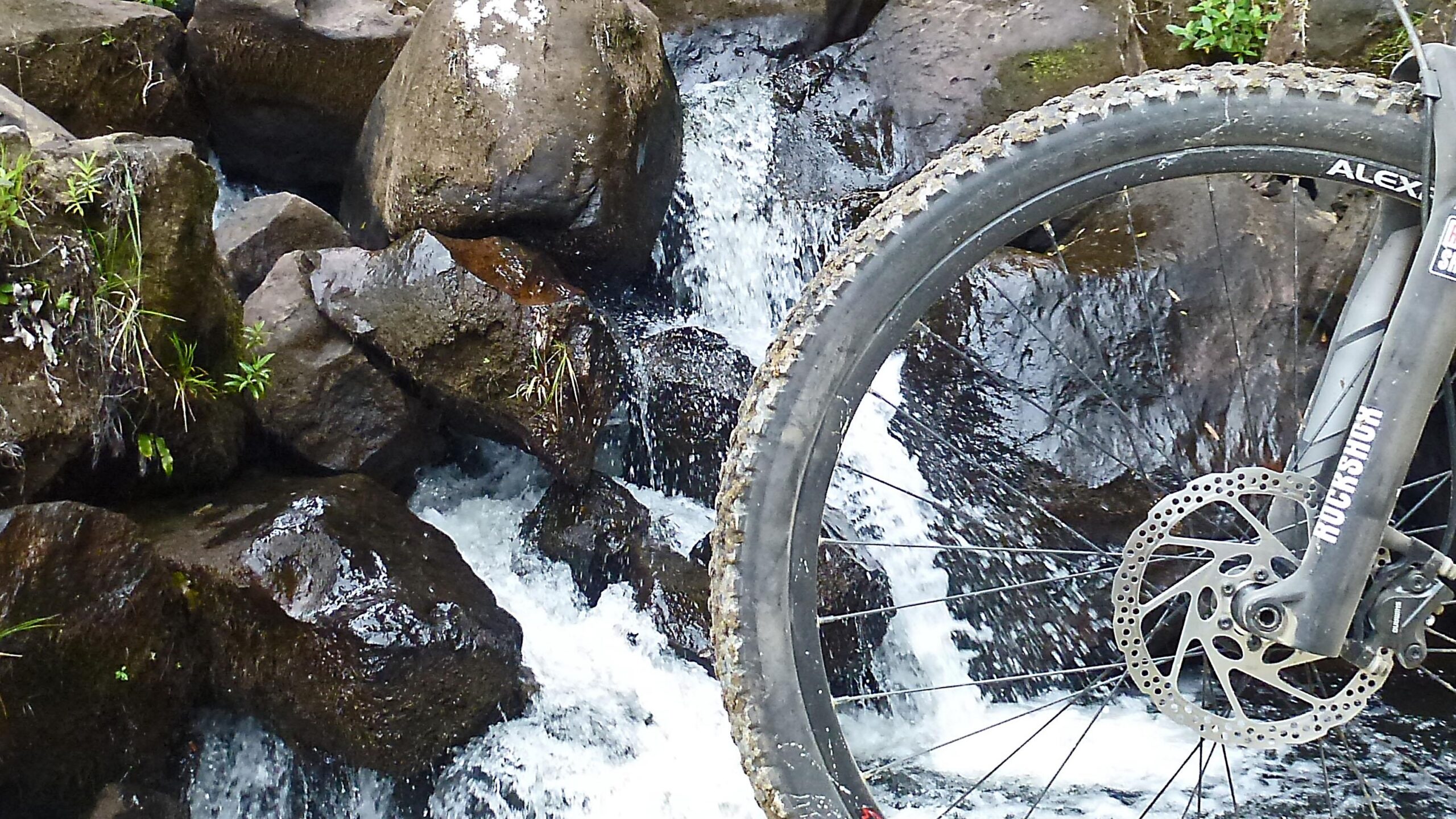 A close-up view of a mountain bike tire alongside a flowing stream, with large rocks and greenery in the background. The bike's disc brake is visible, highlighting its rugged design, while water cascades over the stones. The Timber Trail mountain bike trail.