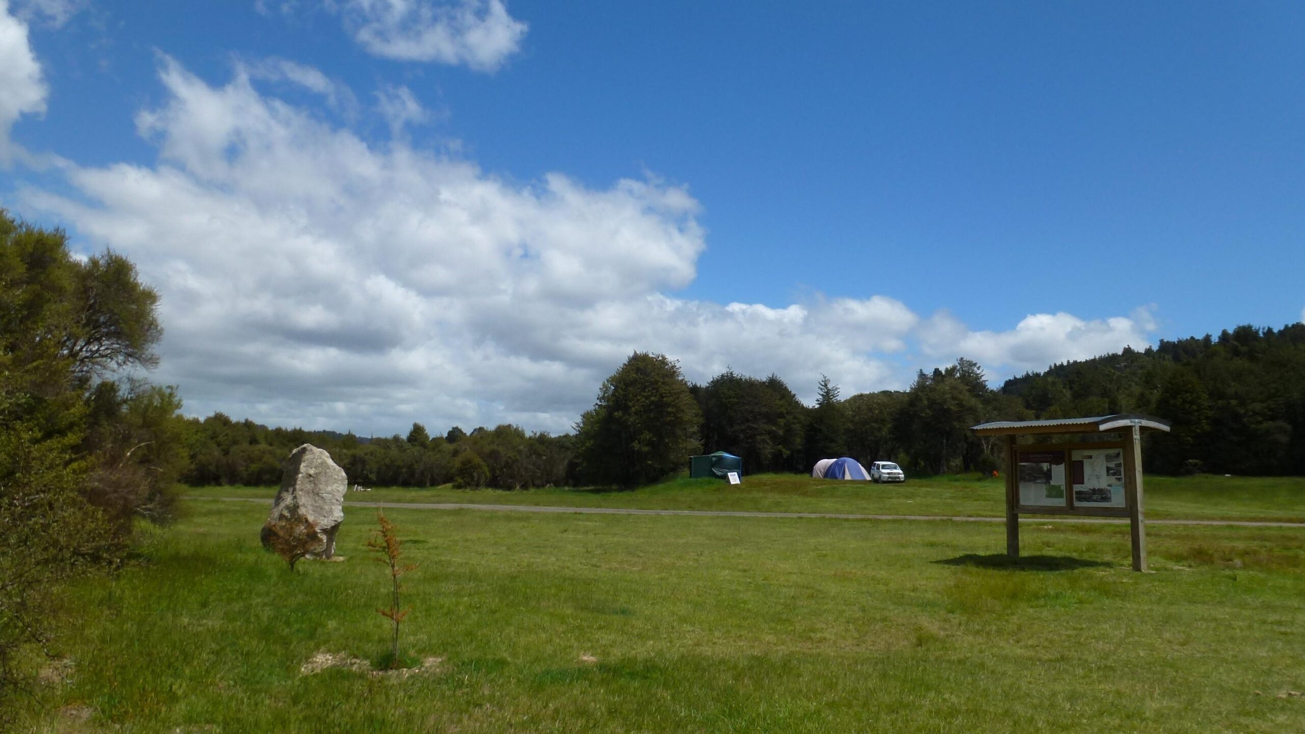 A peaceful outdoor scene featuring a grassy field under a partly cloudy blue sky. In the background, there are two camping tents and a parked vehicle surrounded by trees. A wooden information sign is positioned to the right, providing details about the area. A large rock is visible in the foreground on the left. The Timber Trail mountain bike trail.