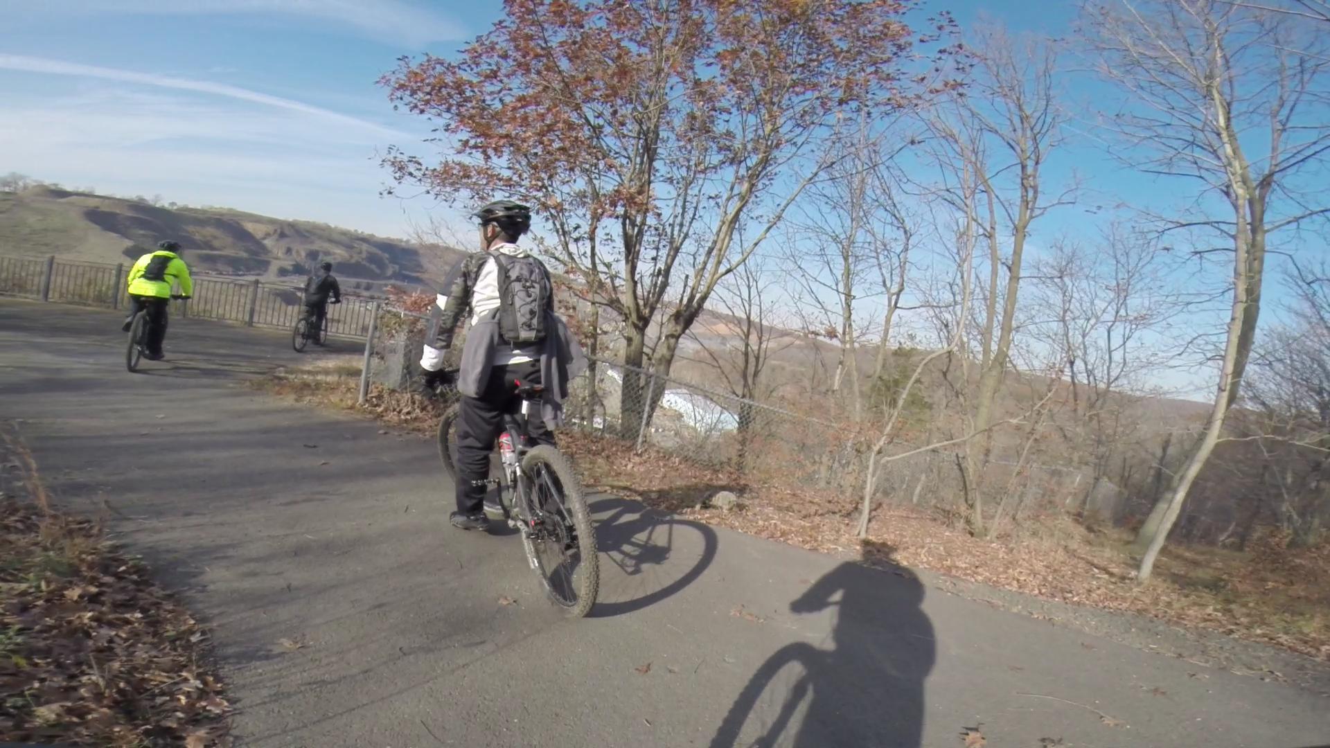 A mountain biker in a black and white outfit rides on a paved trail surrounded by trees with autumn leaves. In the background, another cyclist wearing a bright yellow jacket is seen. The scene captures a clear blue sky and hilly terrain in the distance. Chimney Rock mountain bike trail.
