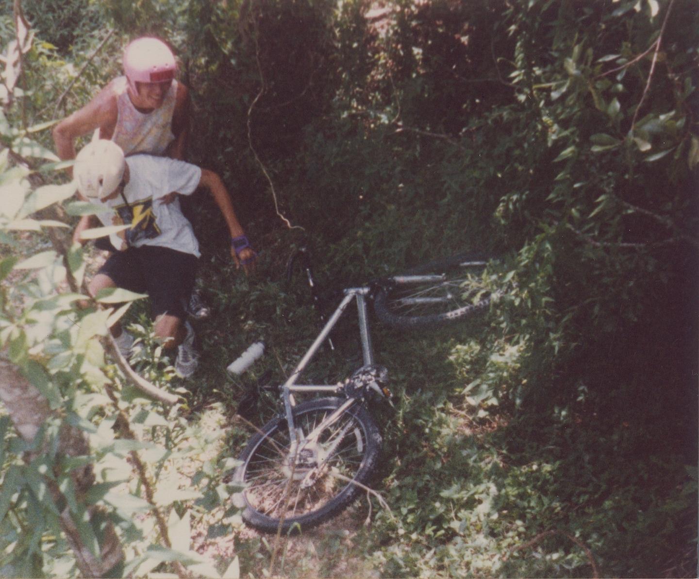 Two individuals in helmets are in a wooded area, one is helping the other who appears to be in a distressed position on the ground near a fallen bicycle. The scene is surrounded by greenery, suggesting a trail or off-road environment. Gran Canyon mountain bike trail.