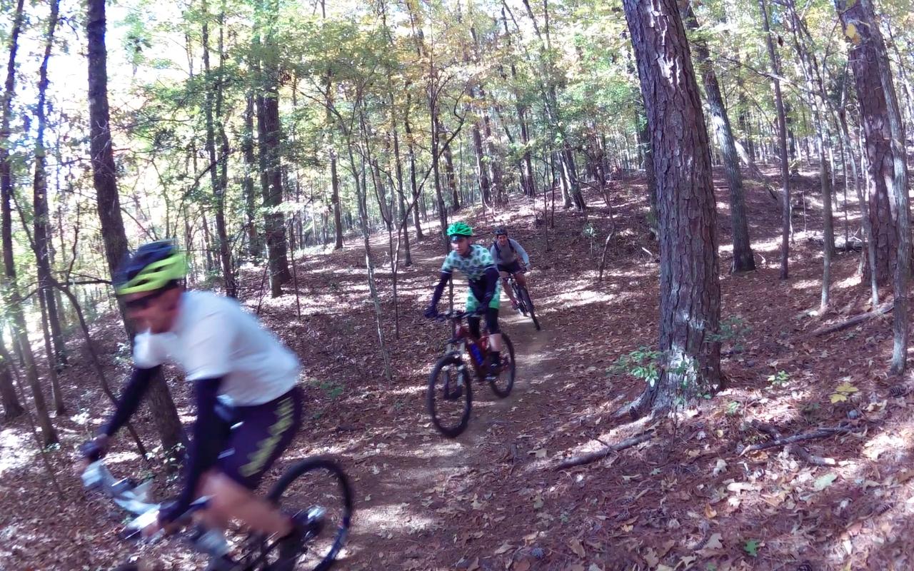 A group of three mountain bikers riding on a dirt trail through a forest with autumn foliage. Two riders are in the foreground, one in a white shirt and the other in a patterned jersey, while the third rider trails behind. The ground is covered in fallen leaves, and tall trees surround the path. Oak Mountain State Park Bump Trail mountain bike trail.