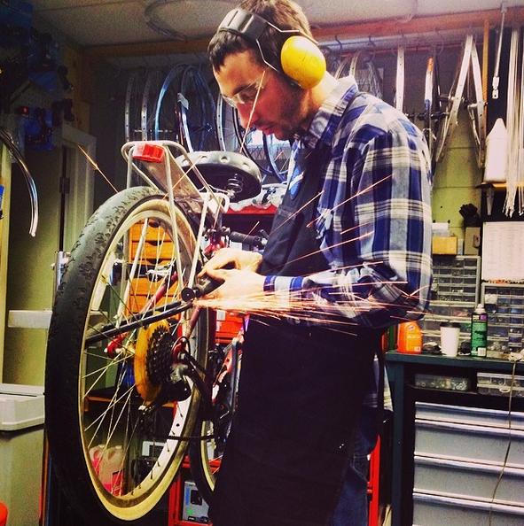 A mechanic wearing protective ear and eye gear works on a bicycle in a workshop, sparks flying from the bike's rear wheel as he tightens components. Various bike parts and tools are visible in the background, illustrating a busy repair environment.