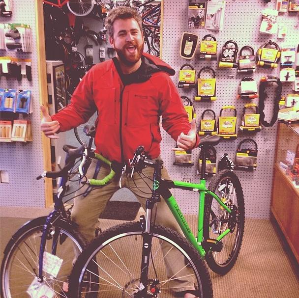 A young man poses playfully between two bicycles in a bike shop, wearing a red jacket and displaying a big grin while giving two thumbs up. The background features various bicycle accessories displayed on a pegboard.