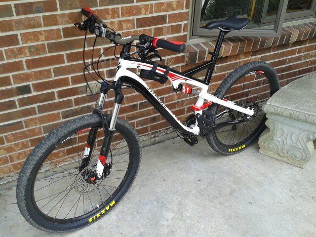 Specialized Camber: A black and white mountain bike with red accents is parked against a brick wall. The bike features thick tires and a suspension system, positioned on a concrete surface beside a stone bench.