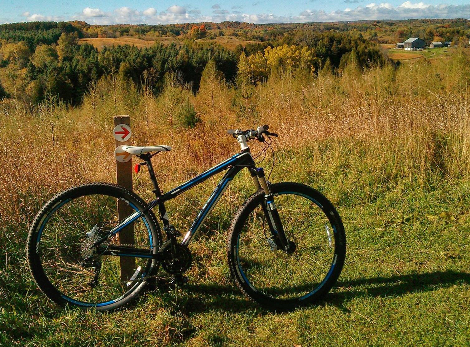 Trek Cobia: A mountain bike is parked next to a trail sign with an arrow, set against a backdrop of rolling hills and trees in autumn colors. The landscape features tall grass and distant buildings under a blue sky with some clouds.