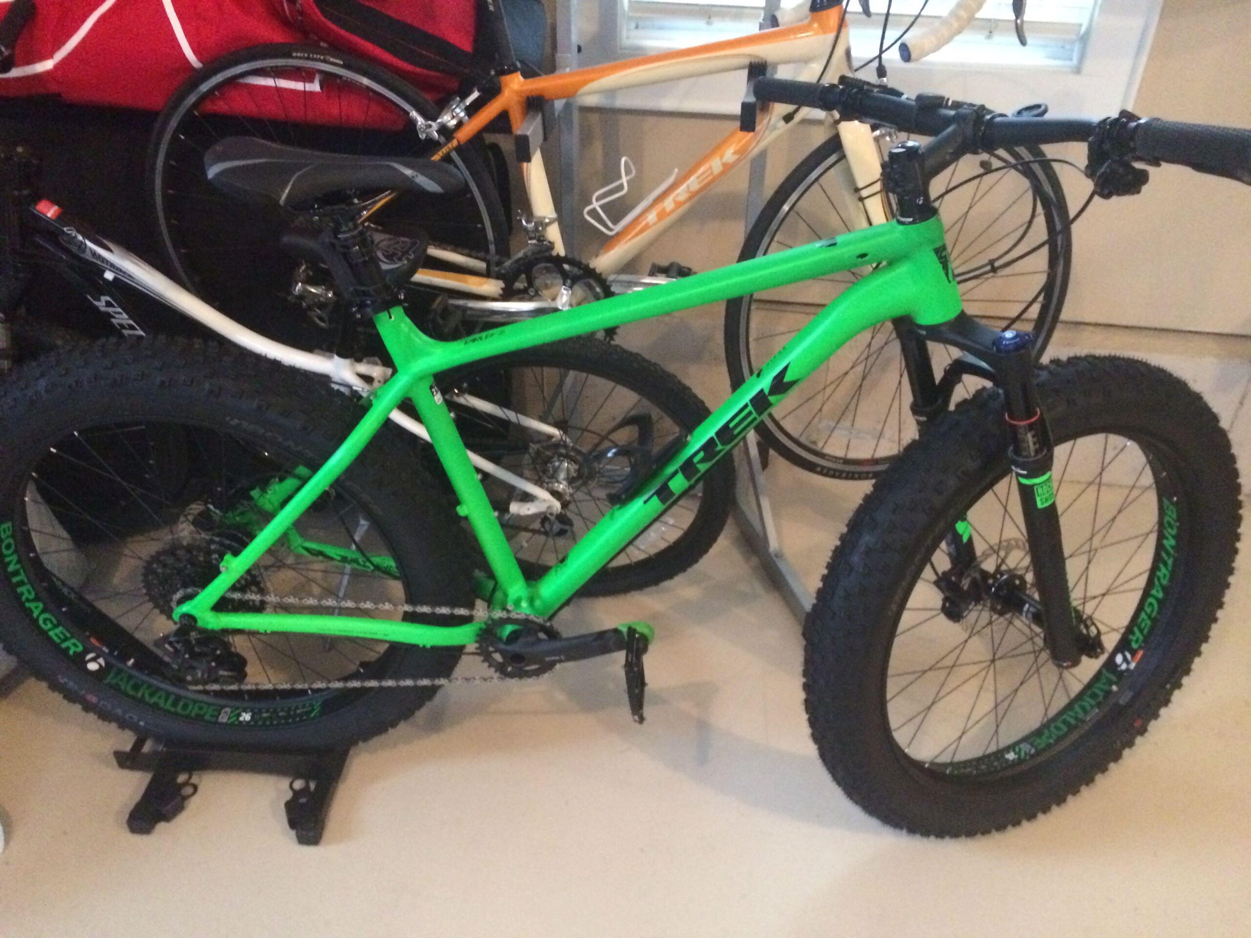 Trek Fuel EX: A bright green Trek mountain bike with fat tires is positioned in a garage alongside a white bike and an orange bike. The bikes are parked on a smooth floor, and the garage has various equipment and tools in the background.