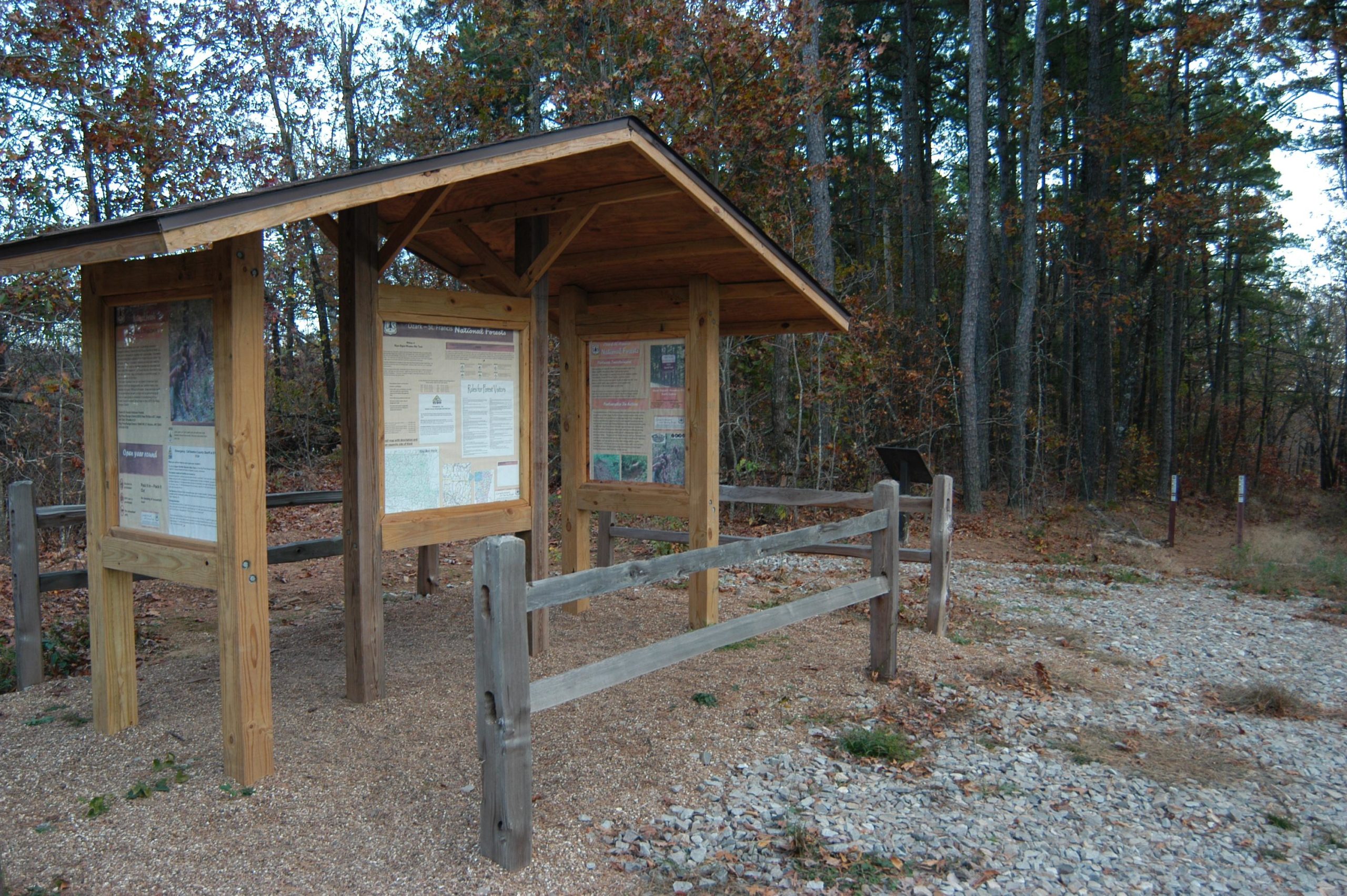 Information kiosk at a trailhead surrounded by trees and fall foliage, featuring educational panels about the area and a gravel path leading into the woods. Upper Buffalo Headwaters Trail System mountain bike trail.