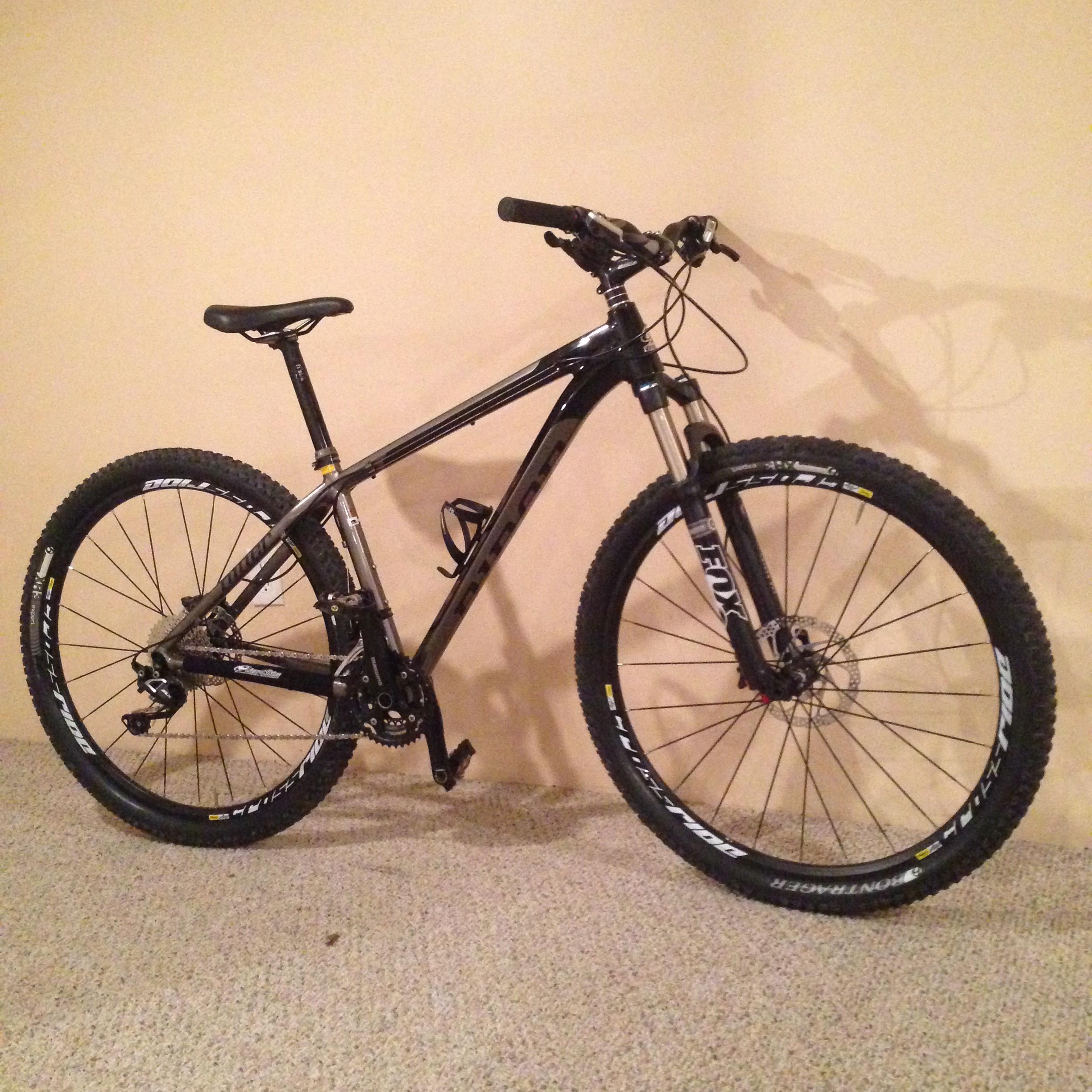 Niner Air 9: A black mountain bike with a lightweight frame and thick, knobby tires, positioned on a carpeted floor against a plain wall. The bike features front suspension, dual disc brakes, and a single gear setup.