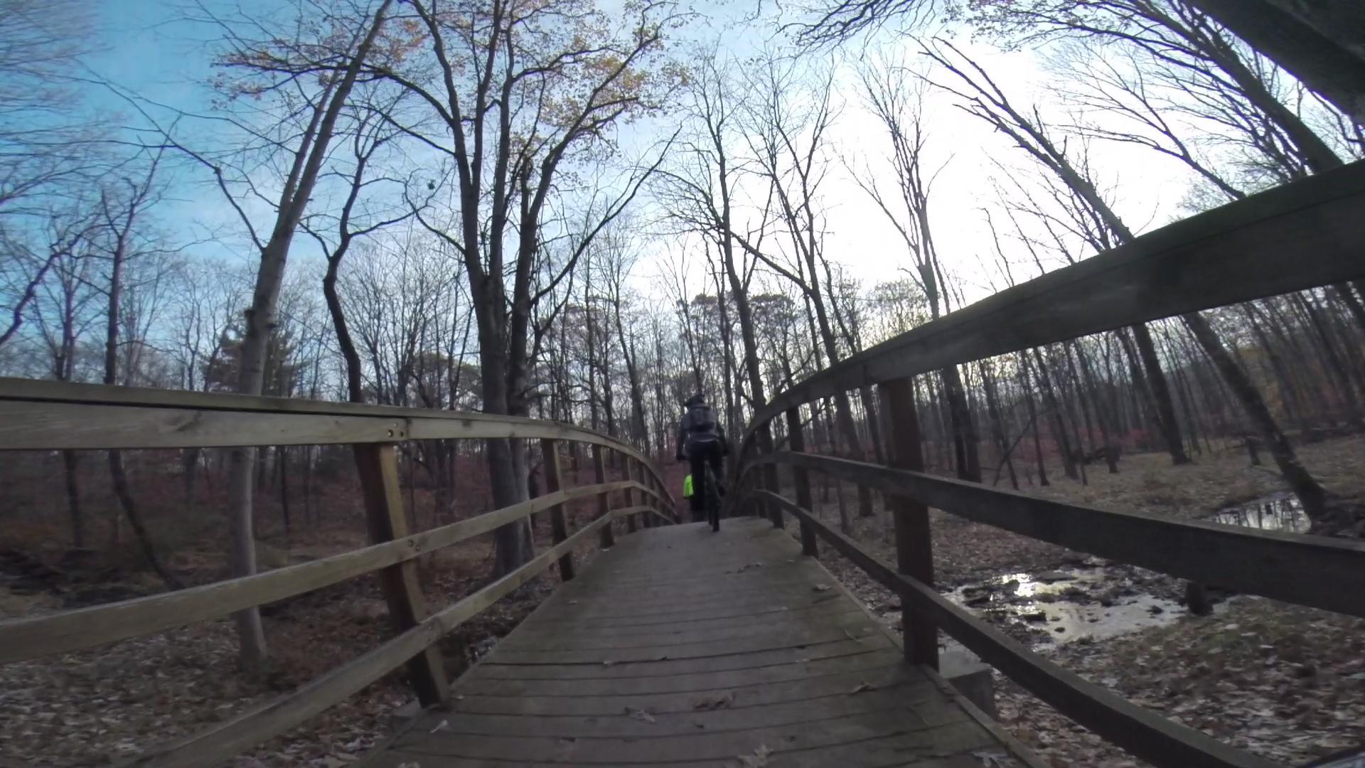 A person riding a bicycle crosses a wooden bridge in a forested area, surrounded by bare trees and a cloudy sky. The ground is covered with fallen leaves, and a small creek can be seen beside the path. Chimney Rock mountain bike trail.