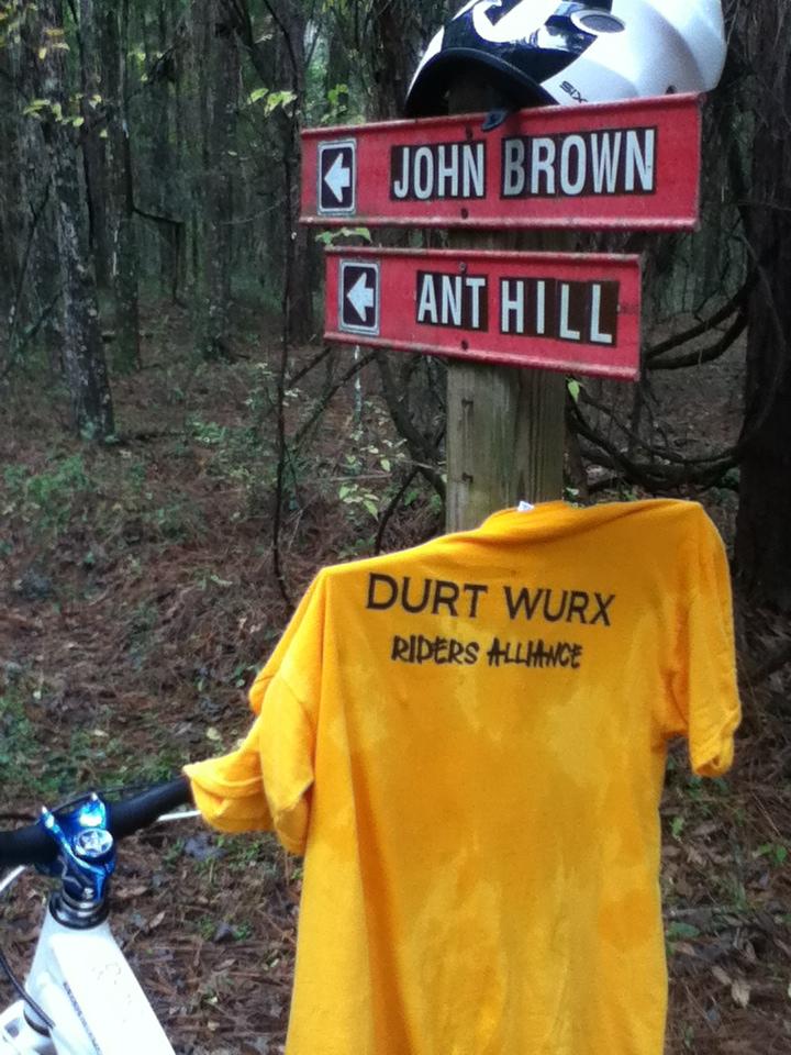 A wooden signpost in a forest displaying directional arrows for "John Brown" and "Ant Hill," with a yellow shirt featuring the text "DURT WURX RIDERS ALLIANCE" draped over a bicycle handlebar. A white helmet rests on top of the signpost. Santos mountain bike trail.