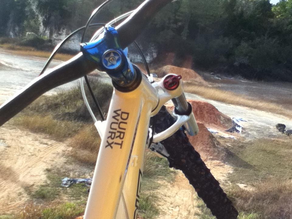 Close-up view of a mountain bike's handlebars and stem, showcasing the brand "DURT WURX." The background features a dirt biking area with trails and mounds of dirt, surrounded by sparse vegetation. Santos mountain bike trail.