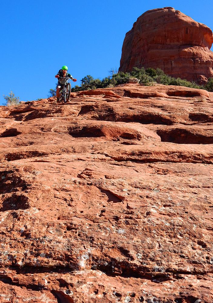 A person in a green helmet rides a unicycle down a steep, rocky terrain with red sandstone, under a clear blue sky. A large rock formation is visible in the background, surrounded by low vegetation. Hangover mountain bike trail.