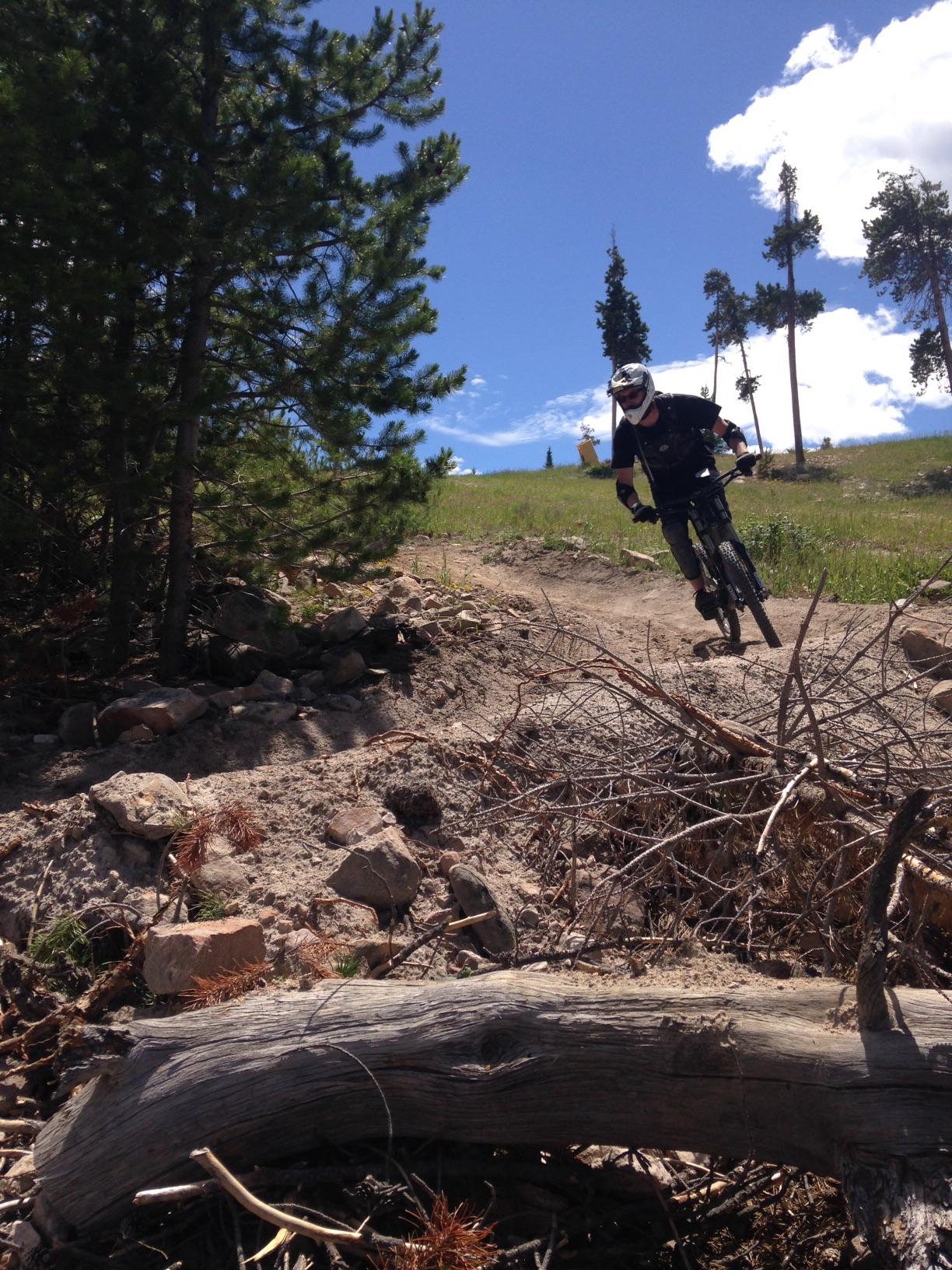 Trek Fuel EX 6 26: A mountain biker navigating a dirt trail surrounded by greenery and trees, with a bright blue sky and scattered clouds in the background. The biker is wearing a helmet and protective gear, and is actively maneuvering past rocks and fallen branches on the trail.